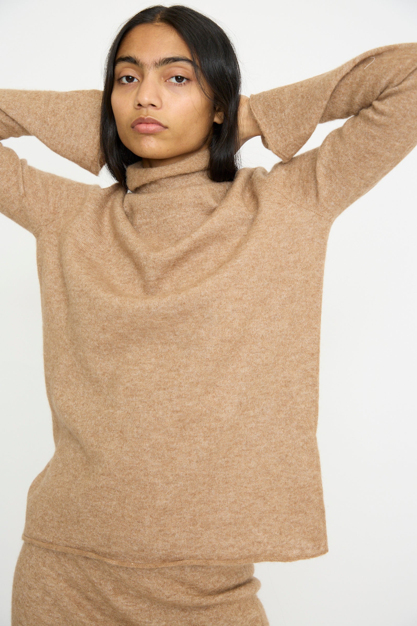 A person in a Lauren Manoogian Funnel Turtleneck in Camel stands poised with arms raised behind their head against a plain background.