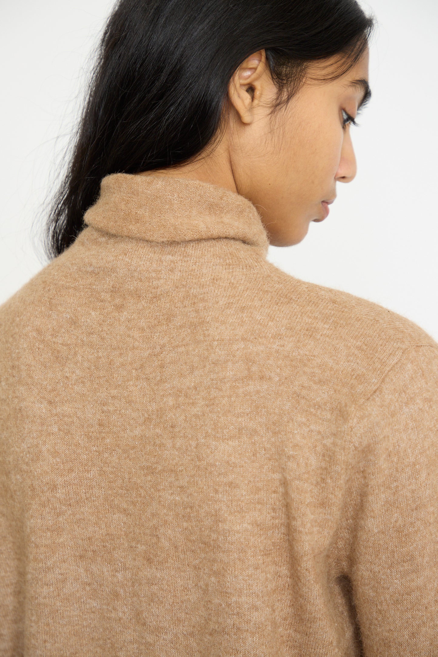 A person in a luxurious Funnel Turtleneck in Camel by Lauren Manoogian faces away from the camera, with dark hair elegantly cascading over the shoulder.