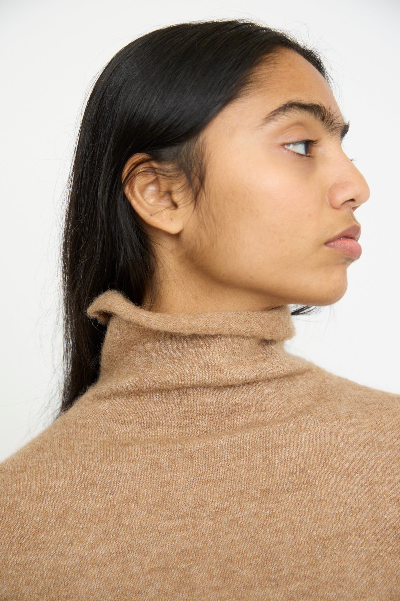 A person with long dark hair wears the Lauren Manoogian Funnel Turtleneck in Camel, crafted from soft baby alpaca wool, viewed in profile against a plain background.