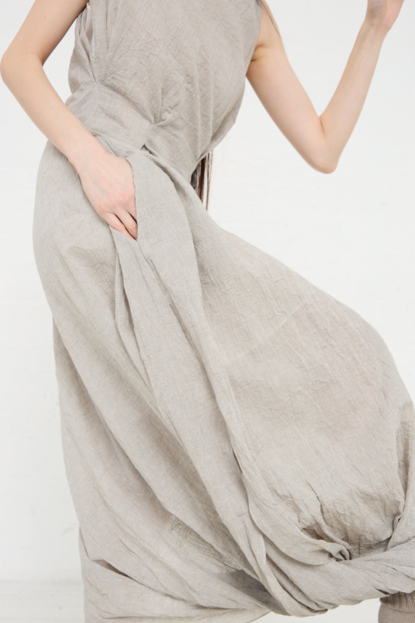 A woman in a beige cotton/linen blend Gauze Twist Dress in Ash by Lauren Manoogian holding the fabric to her side, partial view focusing on the dress and her torso.