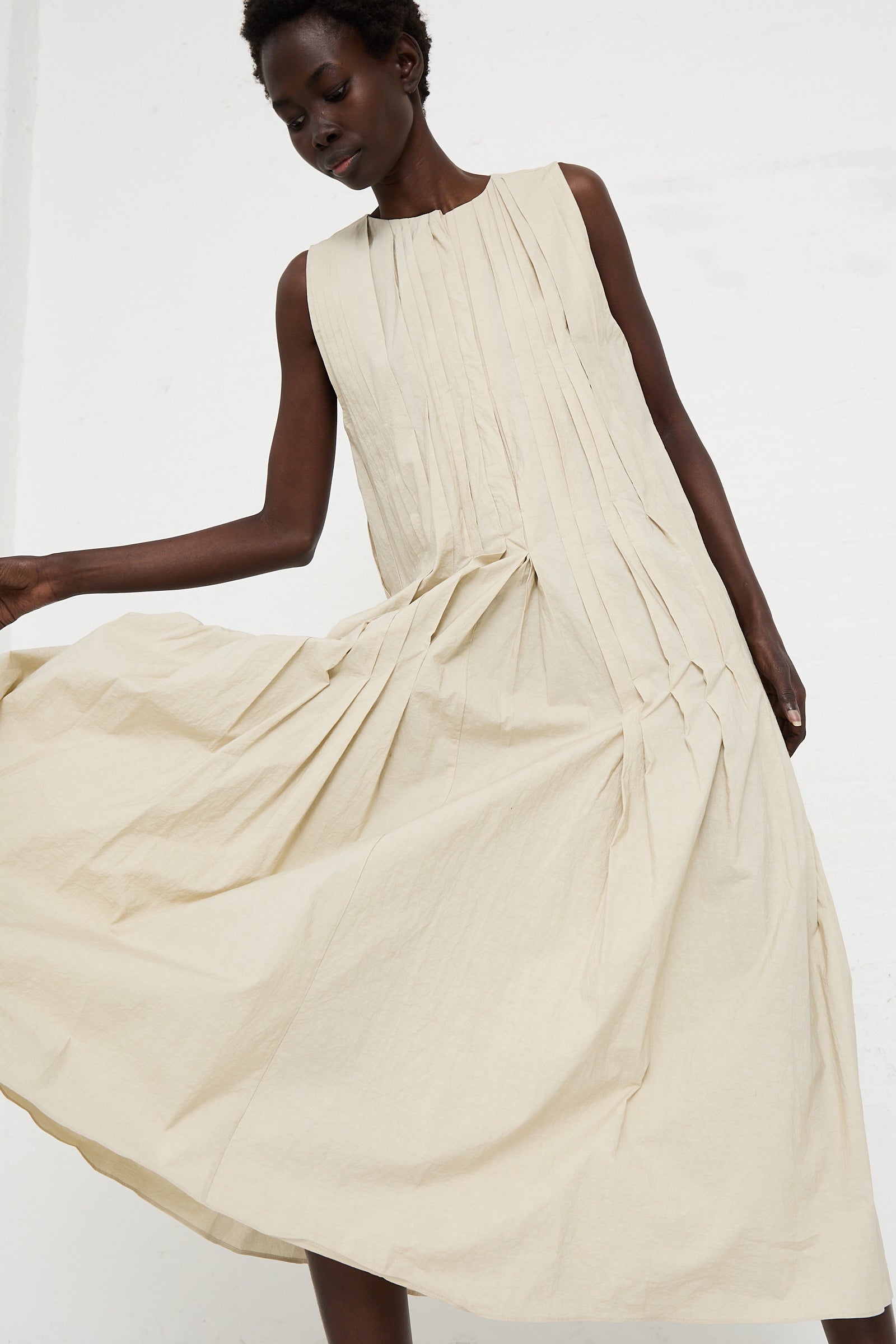 A person wearing the Lauren Manoogian Hand Pleat Dress in Greige crafted from a light-colored cotton/linen blend with pleated details on the front and a full skirt.