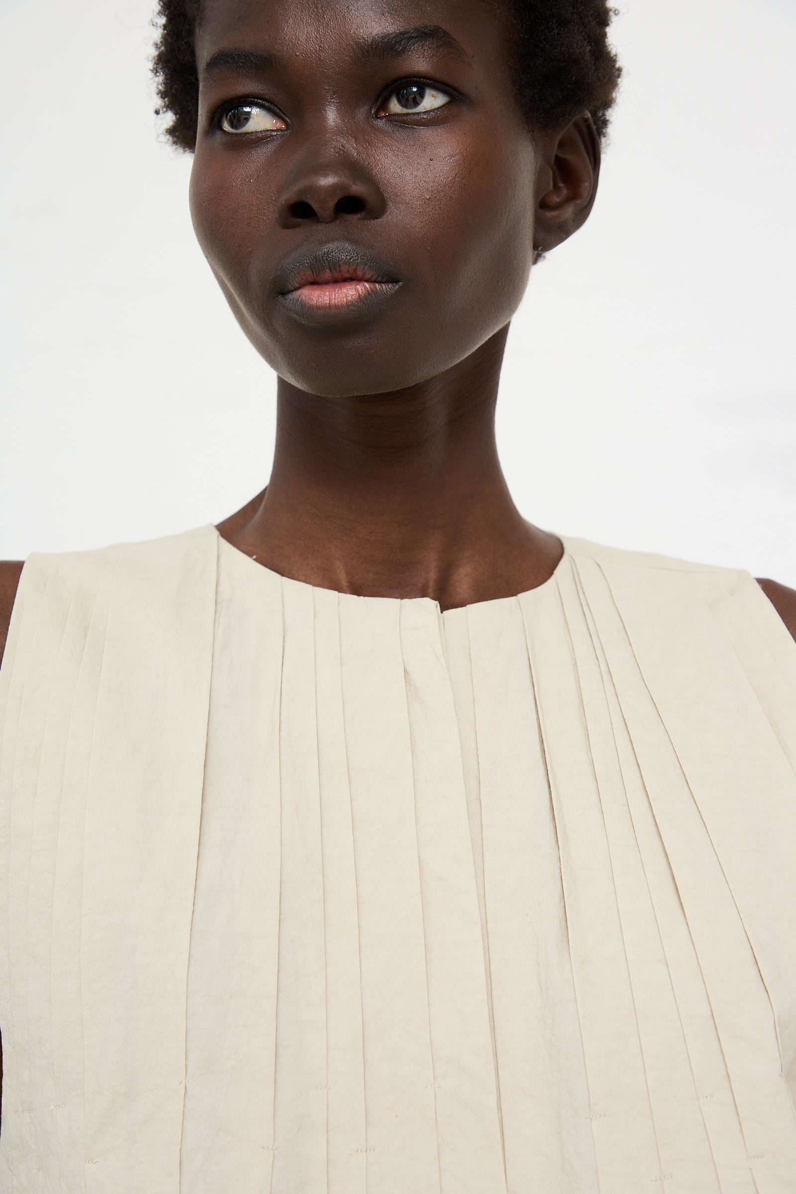 A person with short hair looks slightly to the side, wearing a Hand Pleat Dress in Greige by Lauren Manoogian against a plain background.