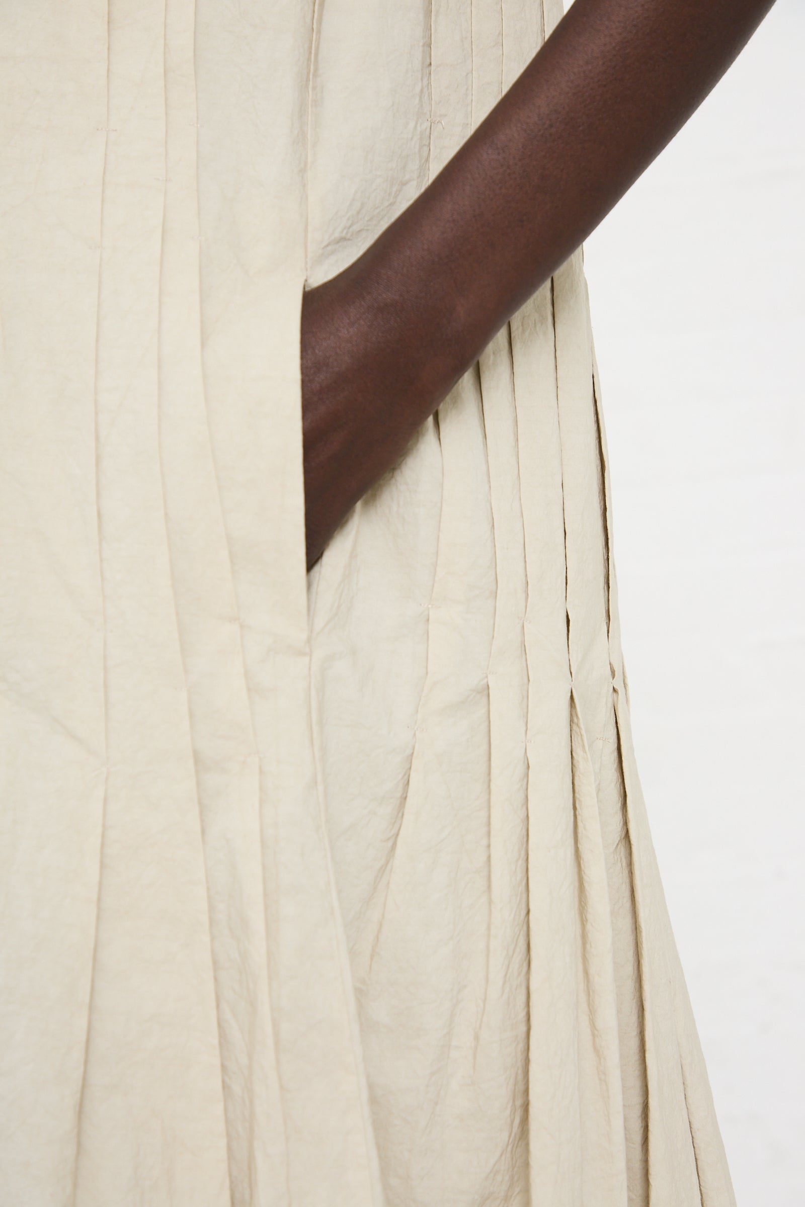 Close-up of a person with a dark complexion wearing a light beige Hand Pleat Dress in Greige by Lauren Manoogian, showing their hand casually placed in a pocket.