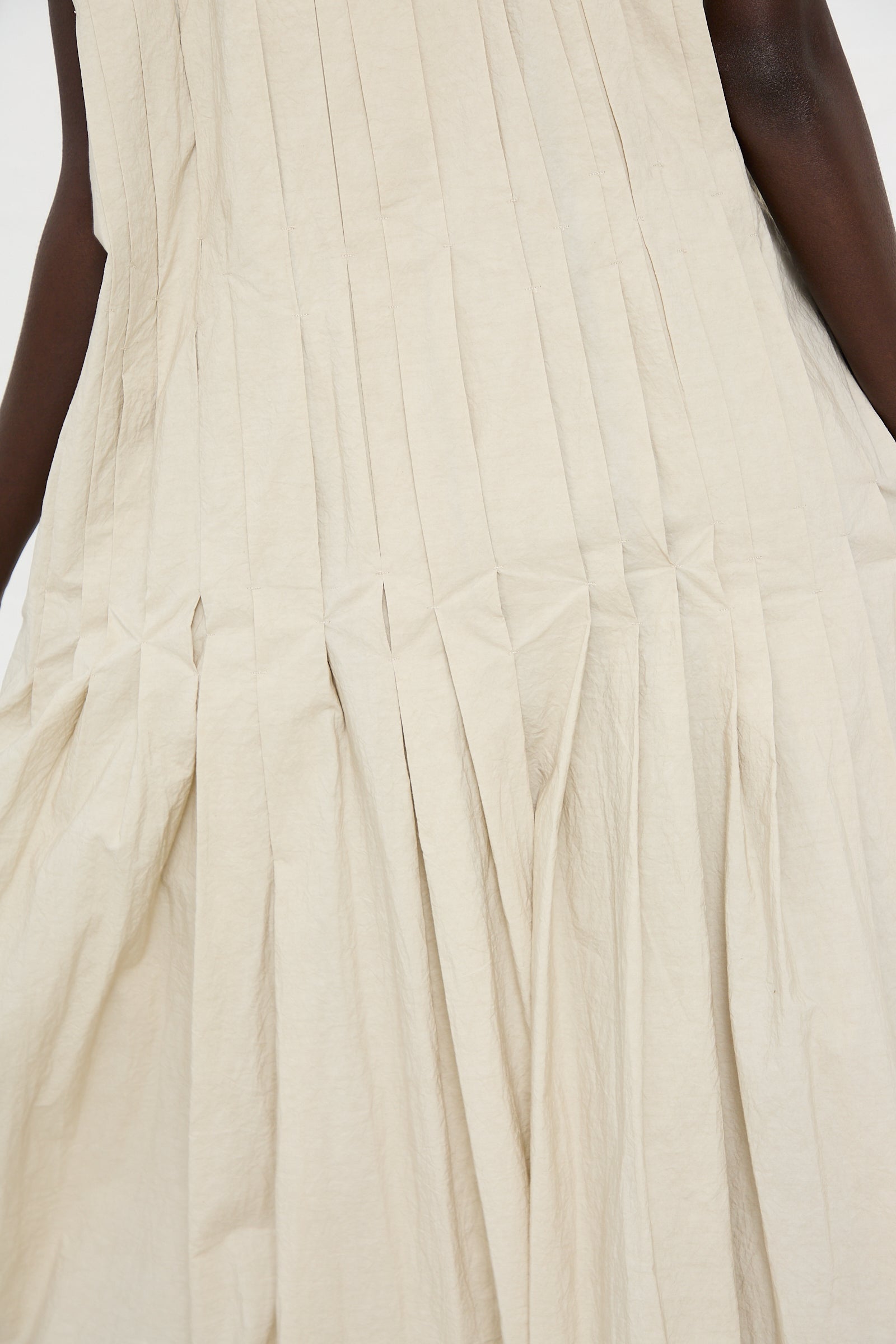 Close-up of a person wearing the Hand Pleat Dress in Greige by Lauren Manoogian, a beige, pleated, and textured sleeveless maxi dress made from hand-dyed cotton/linen blend fabric.