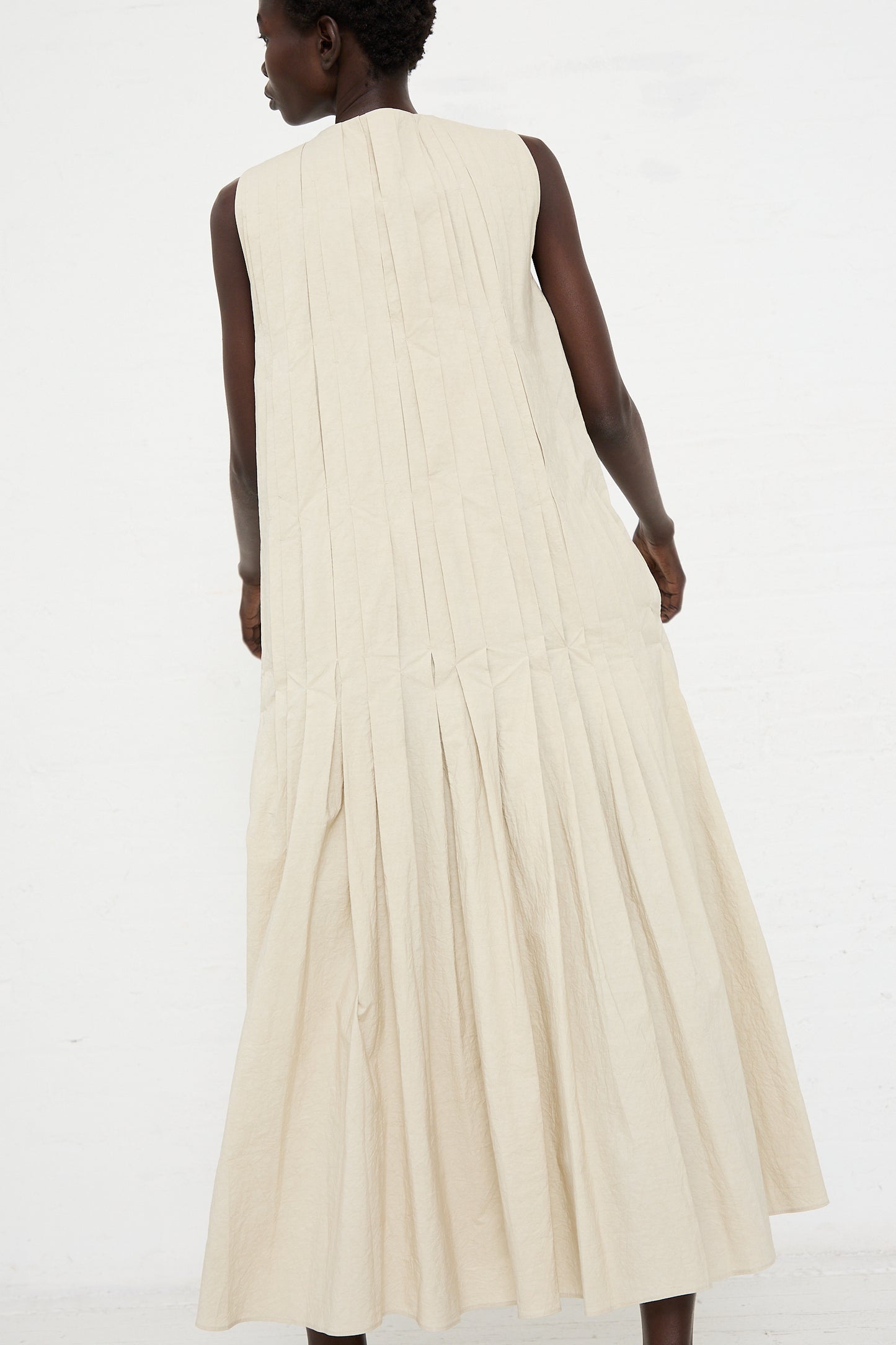 A person is facing away from the camera, wearing a Hand Pleat Dress in Greige by Lauren Manoogian made of a hand dyed cotton/linen blend fabric that extends to their ankles. The background is a plain white wall.