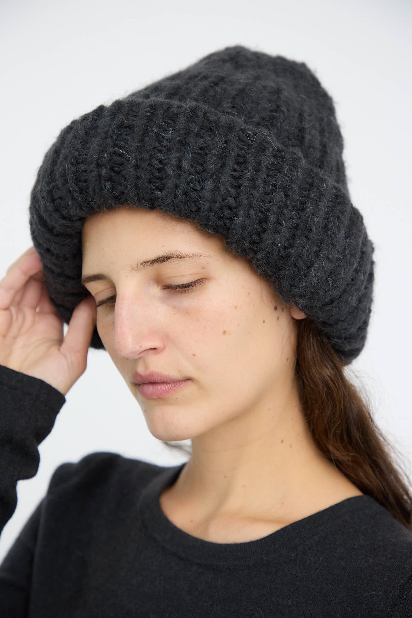 A person wearing the Lauren Manoogian Handknit Big Hat in Ink, crafted from an alpaca/wool blend and made in Peru, adjusts it with one hand. Their eyes are closed, and they are wrapped snugly in a dark sweater.