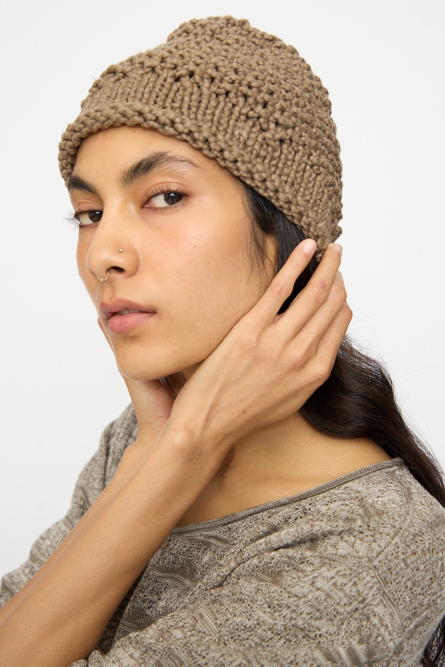 A person wearing the Lauren Manoogian Handknit Crepe Hat in Bark and a patterned top looks at the camera, touching the side of their head.