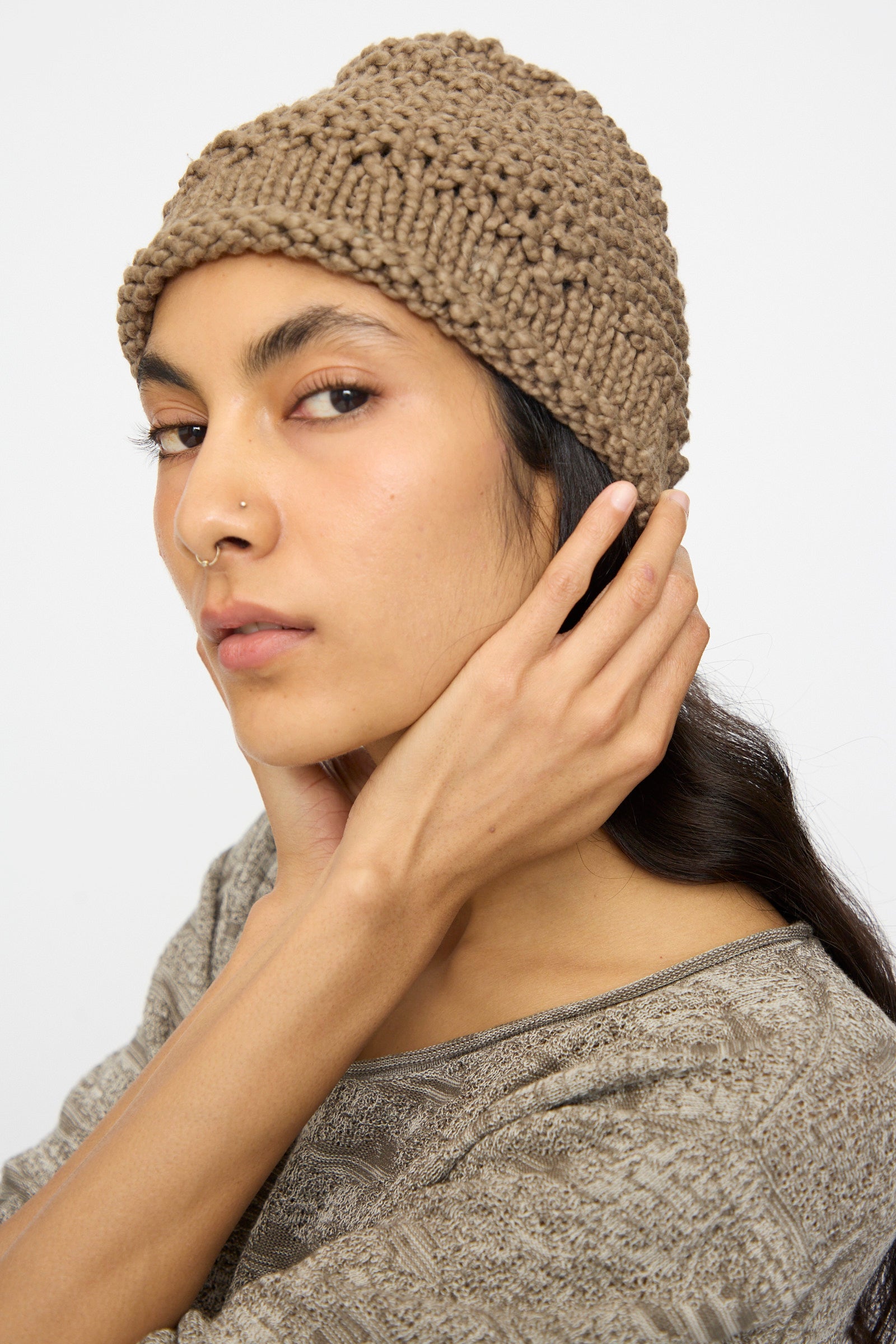 A person wearing the Lauren Manoogian Handknit Crepe Hat in Bark and a patterned top looks at the camera, touching the side of their head.