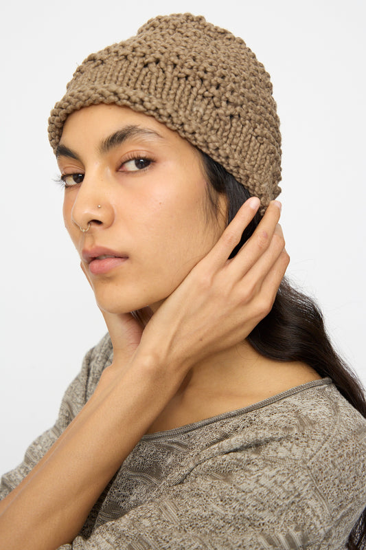 A person wearing the Lauren Manoogian Handknit Crepe Hat in Bark and a patterned top looks at the camera, touching the side of their head.