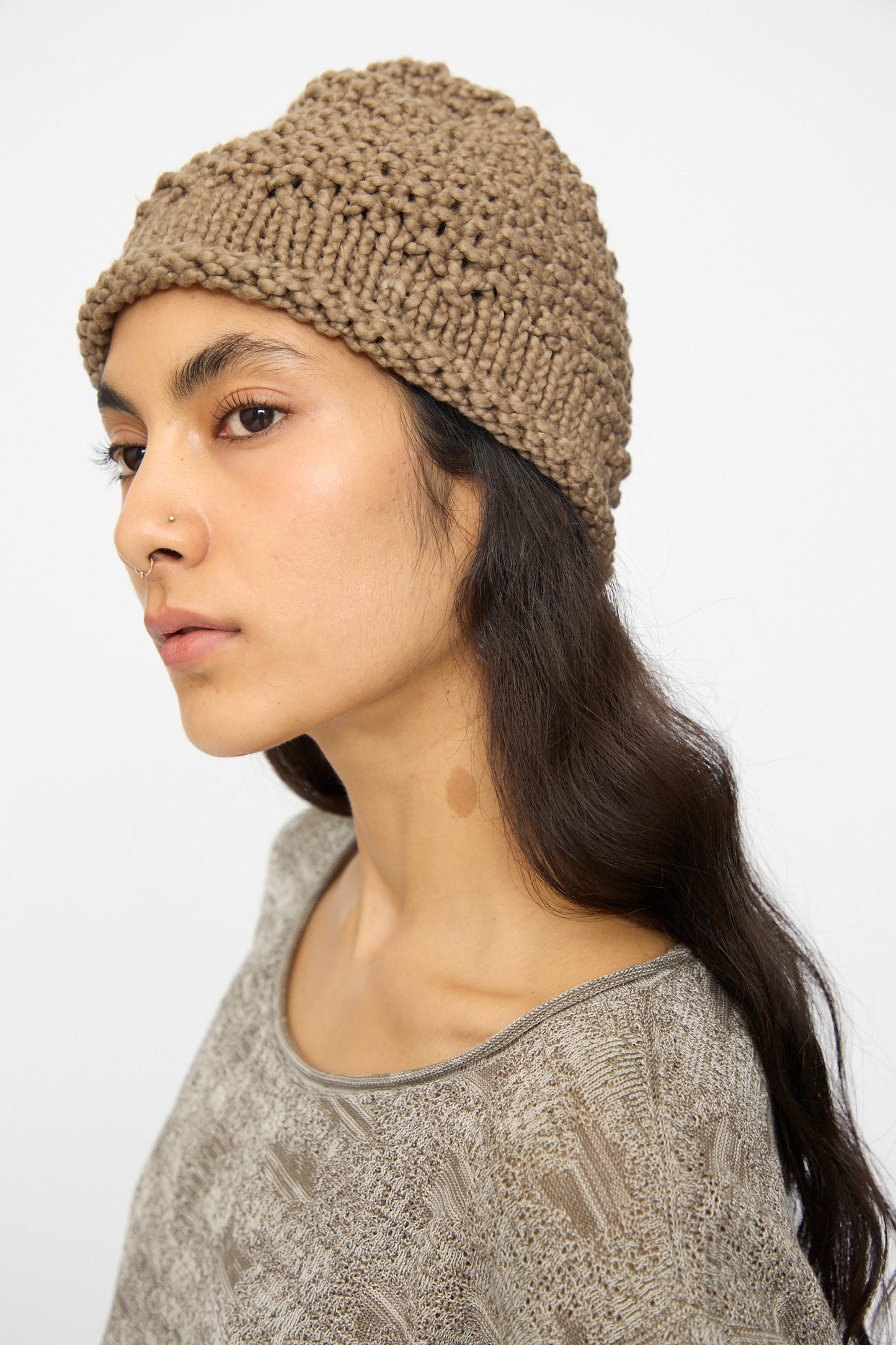 A person wears the Lauren Manoogian Handknit Crepe Hat in Bark and a beige patterned top, looking to the side against a plain light background.