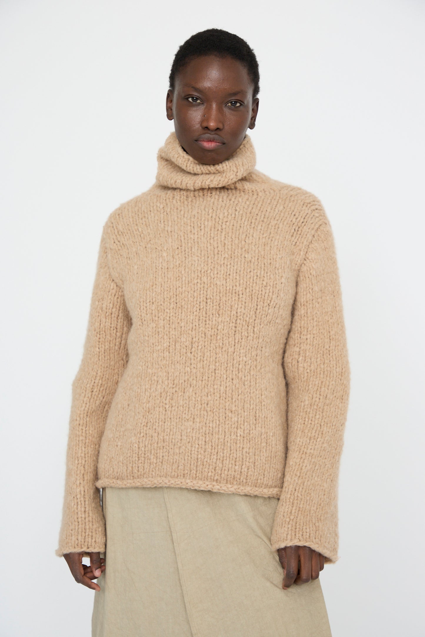 A person wears the Lauren Manoogian Handknit Funnel Sweater in Camel, paired with a tan skirt, standing against a plain white background.