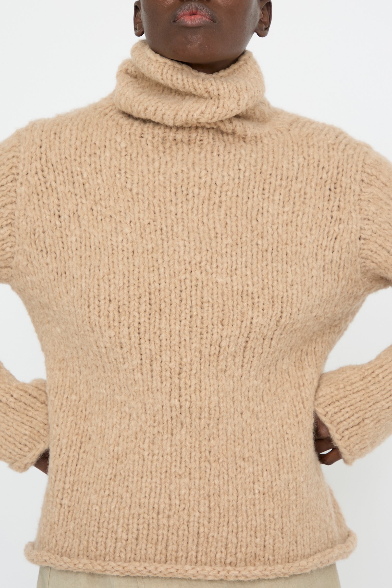 A person with hands on hips wearing a Lauren Manoogian Handknit Funnel Sweater in Camel, set against a plain background.