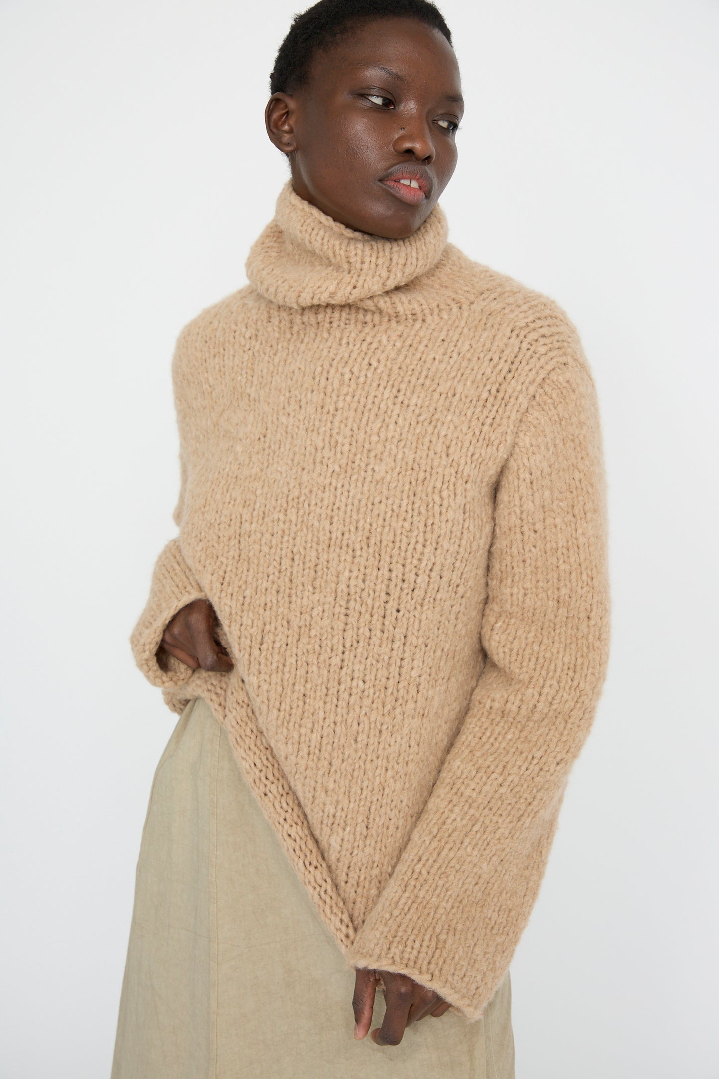 A person wears the Lauren Manoogian Handknit Funnel Sweater in Camel paired with a light skirt, posing against a plain background.