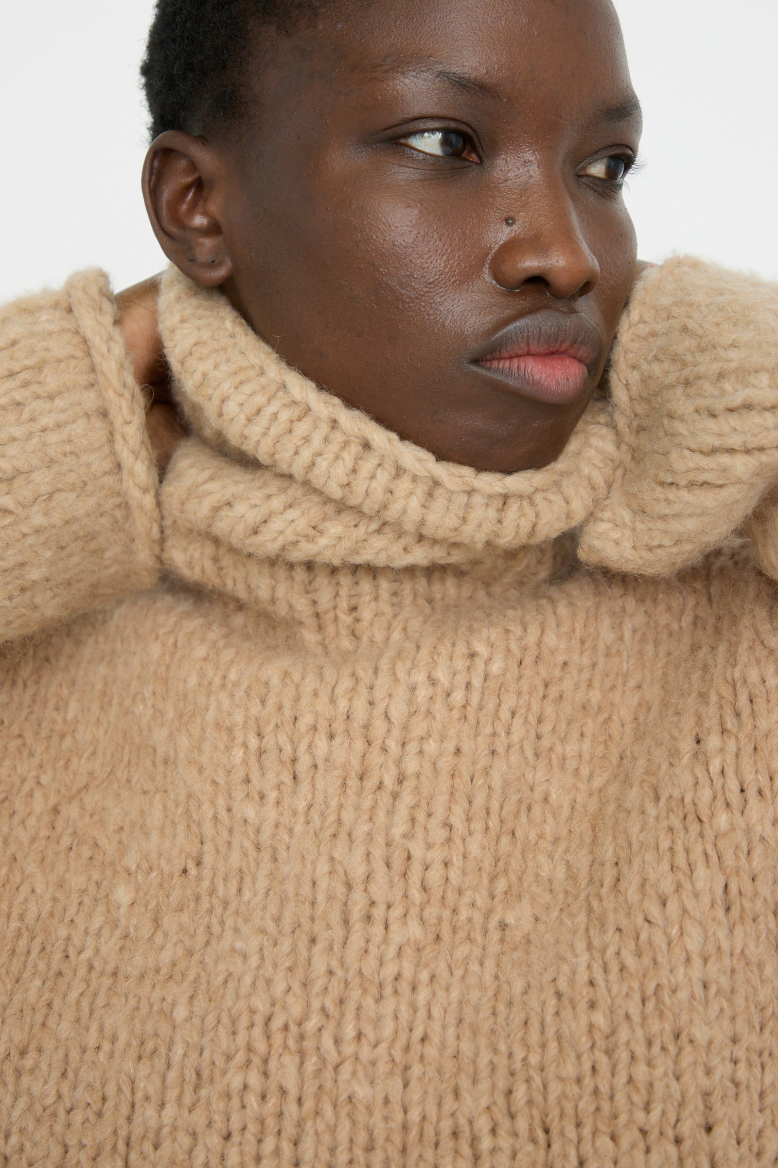 A close-up of a model gazing to the side, wearing a Lauren Manoogian Handknit Funnel Sweater in Camel.