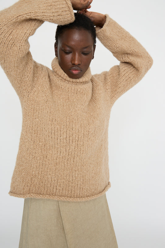 A person in a Lauren Manoogian Handknit Funnel Sweater in Camel raises their arms against a plain background.