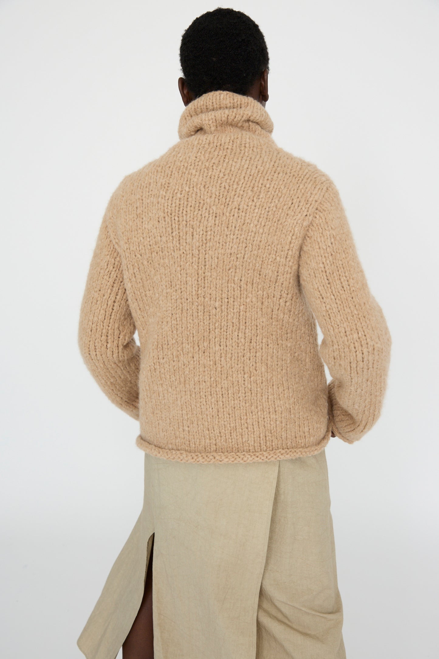 A person stands against a plain background, wearing Lauren Manoogian's Handknit Funnel Sweater in camel, paired with a beige skirt featuring a back slit.