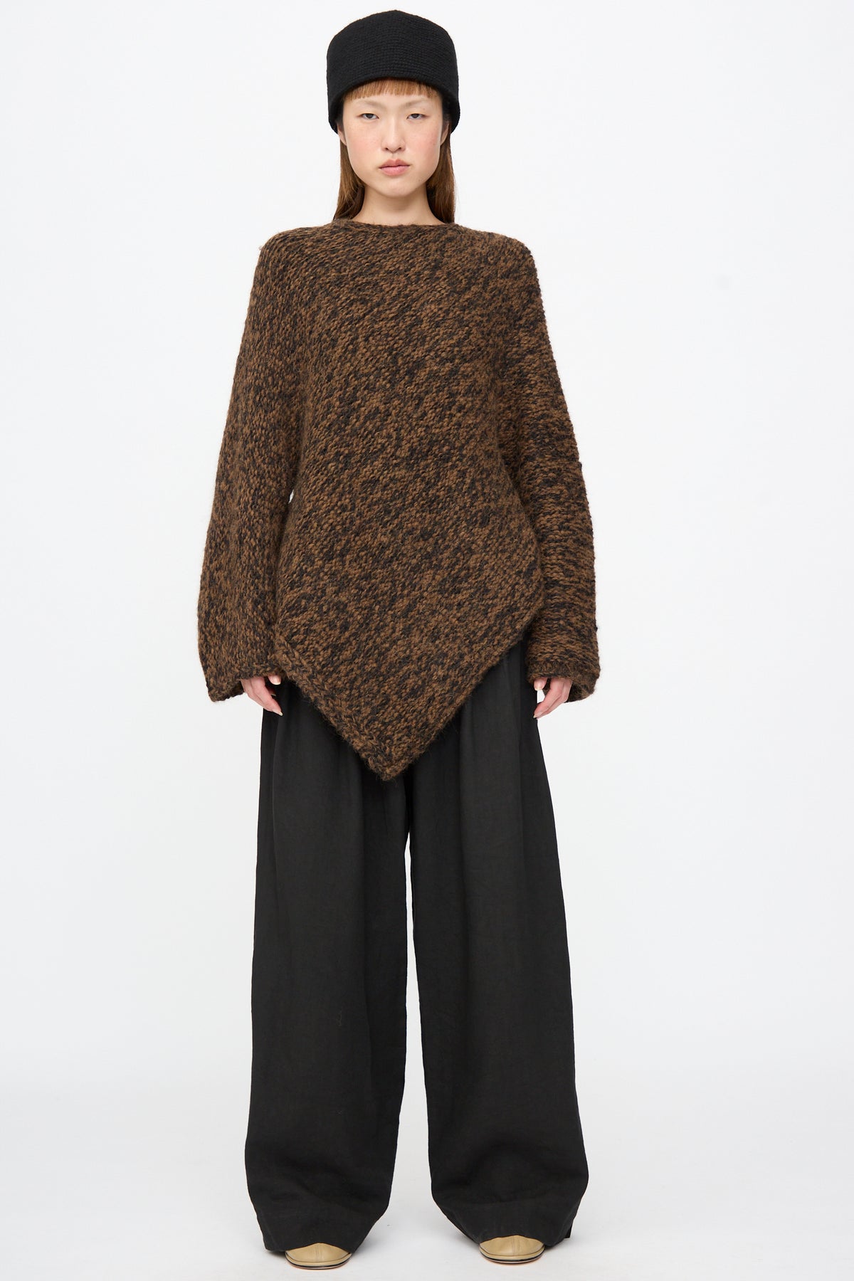 A person wearing a relaxed fit, rust marl Handknit Manta Pullover with a pointed asymmetric hem and wide-leg black trousers, paired with a black hat.