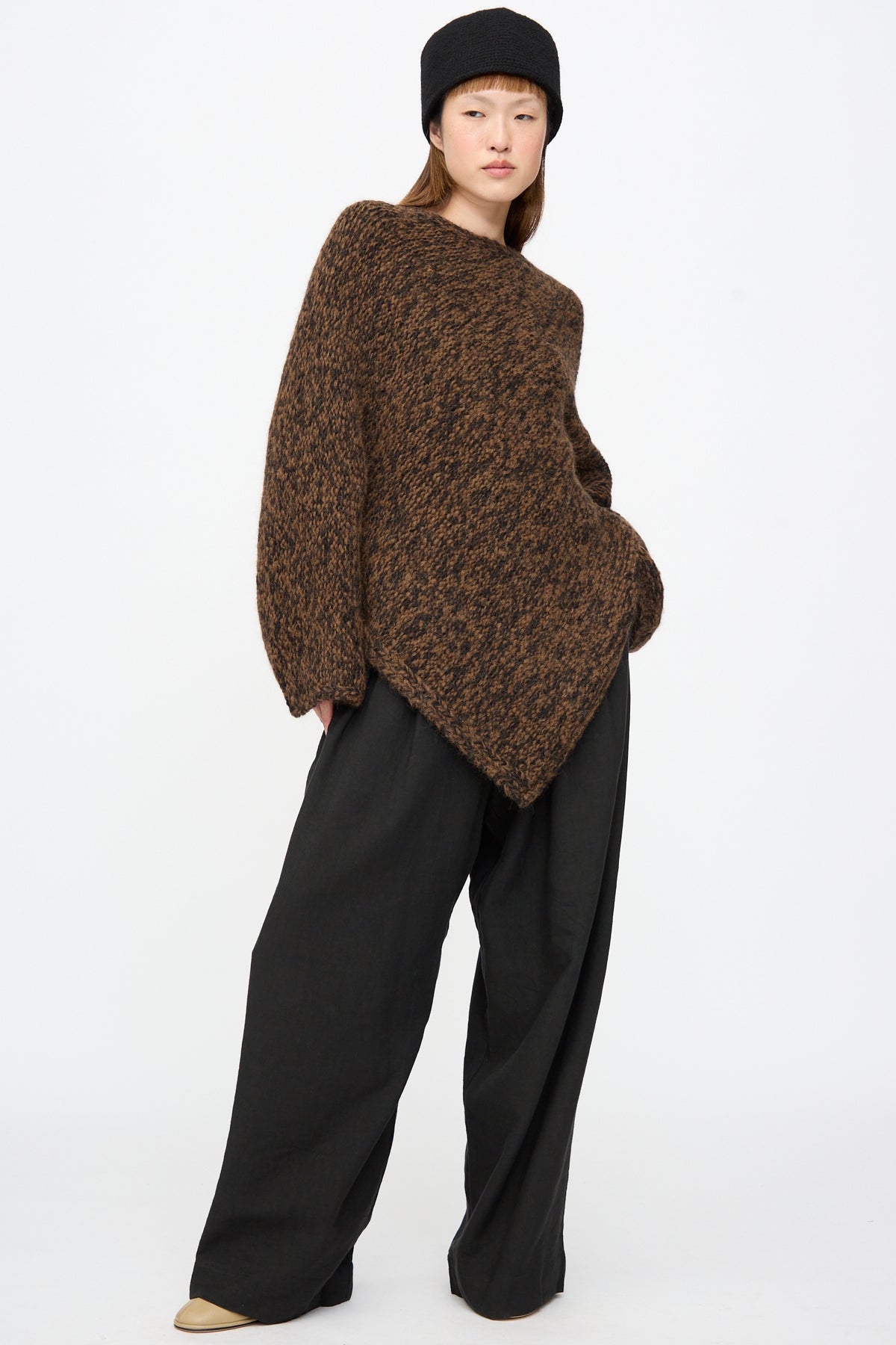 A person wearing a brown chunky alpaca Lauren Manoogian Handknit Manta Pullover in Rust Marl with an asymmetrical hem, black wide-leg trousers, and a black hat.