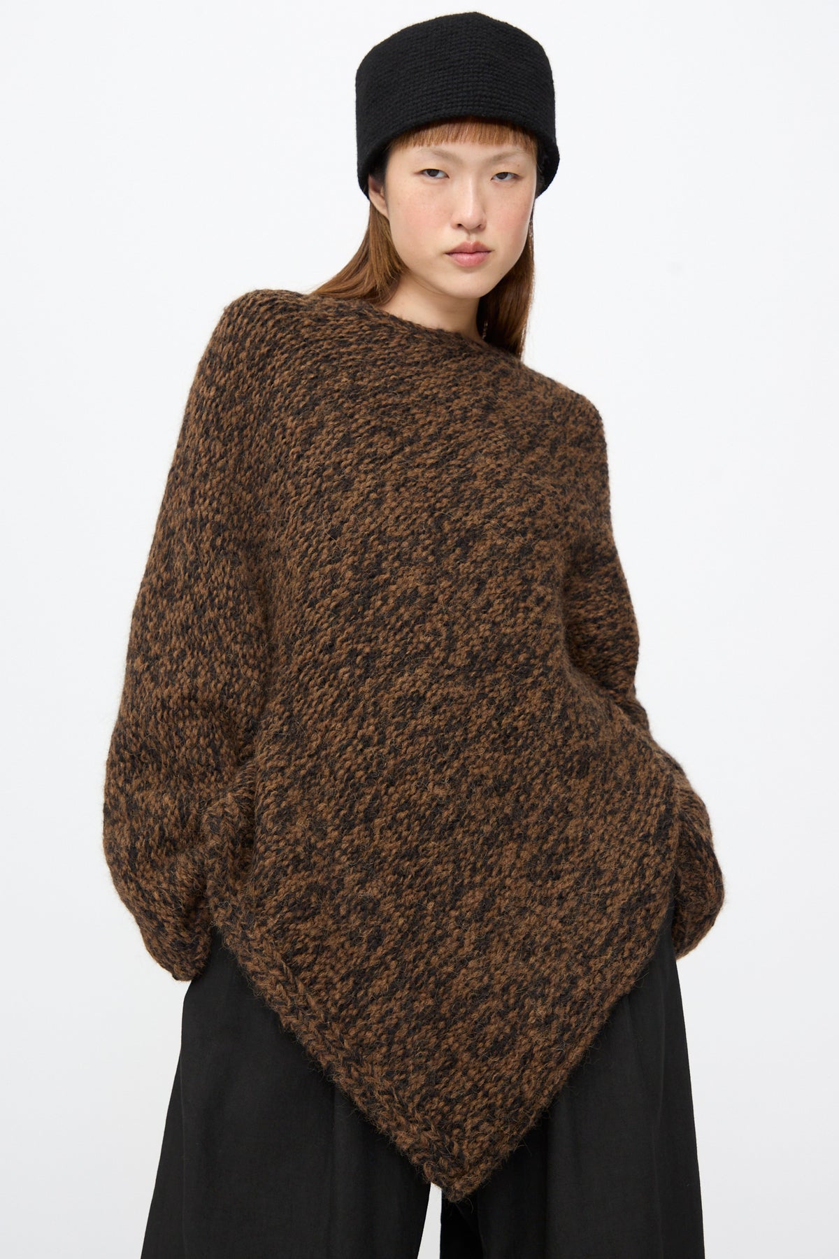 A person wearing a brown and black speckled Handknit Manta Pullover in Rust Marl with an asymmetrical hem and a black beanie.