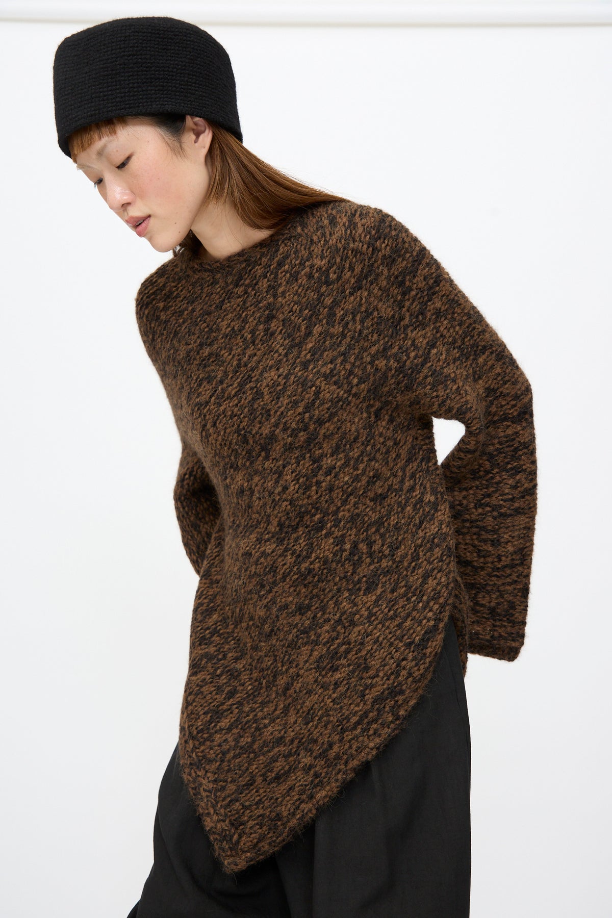 A person wearing a rust marl Lauren Manoogian Handknit Manta Pullover with a chunky texture and asymmetrical hem, paired with a black hat.