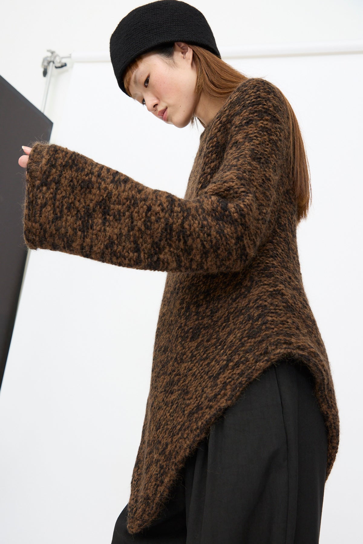 A person wearing a textured, dark brown, chunky Handknit Manta Pullover in Rust Marl with an asymmetrical hem and a black hat.