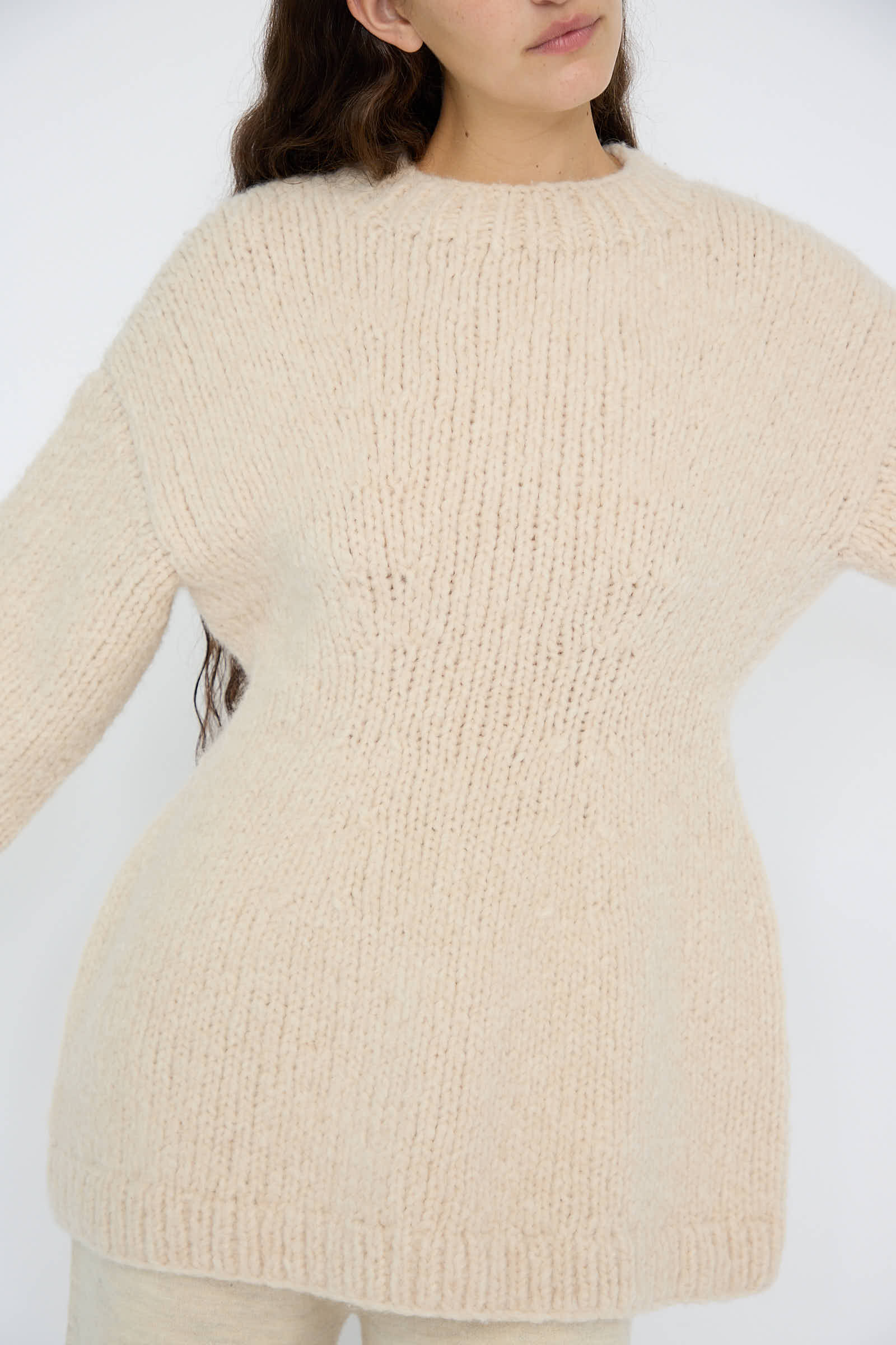 A person wearing the Handknit Peplum Pullover in Alabaster by Lauren Manoogian, made from luxurious baby alpaca wool, is posing against a plain white background.