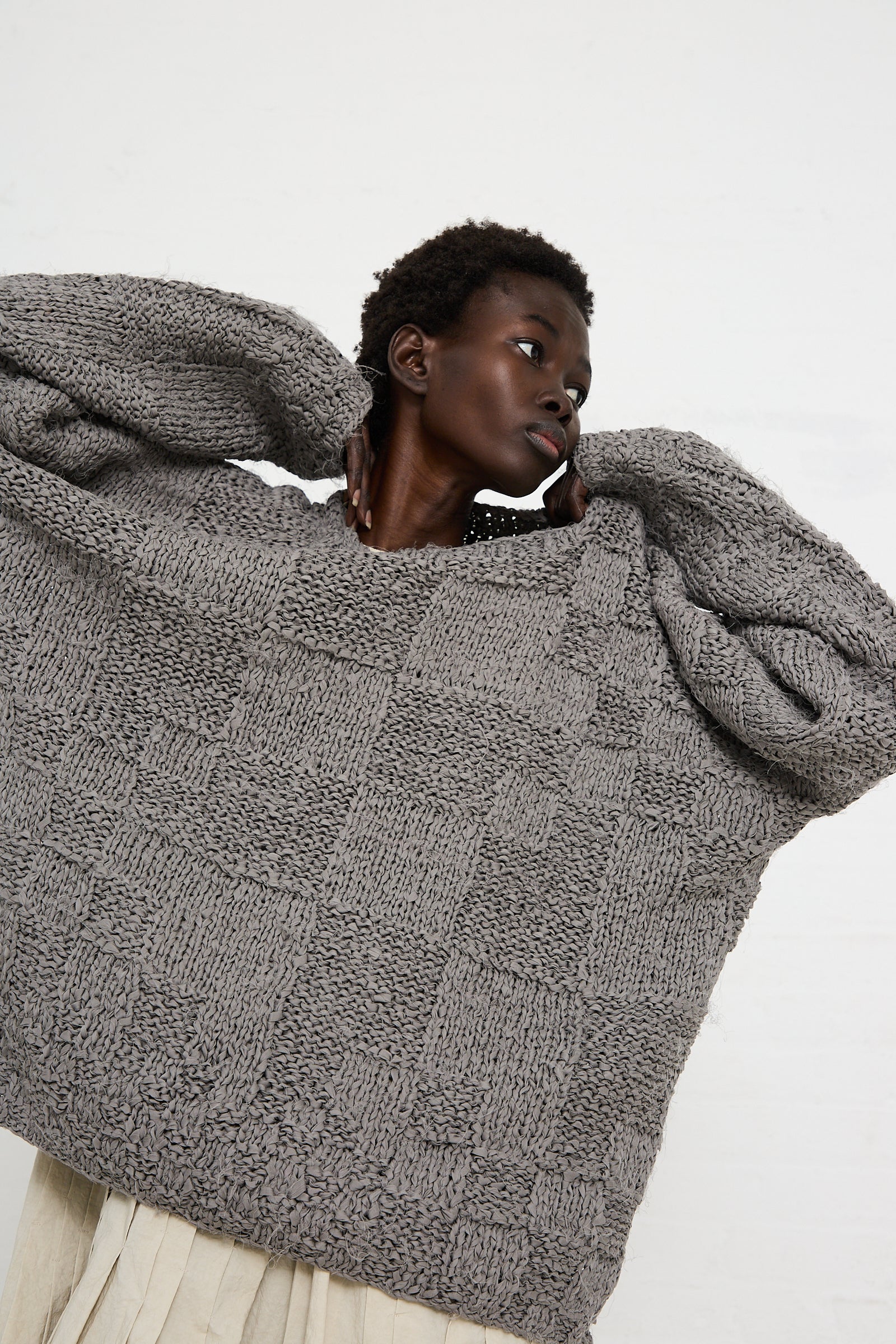 Person wearing a Handknit Pullover Sweater in Gris by Lauren Manoogian with a relaxed fit and textured pattern, paired with an off-white skirt, poses with arms lifted against a plain white background.