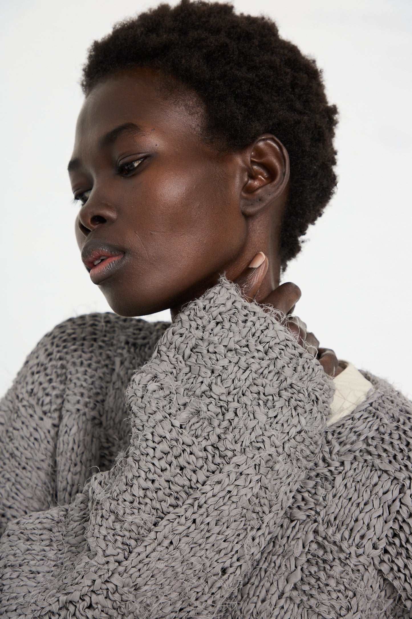 A person with short curly hair wearing a gray Handknit Pullover Sweater in Gris by Lauren Manoogian looks down while gently touching their neck with one hand.