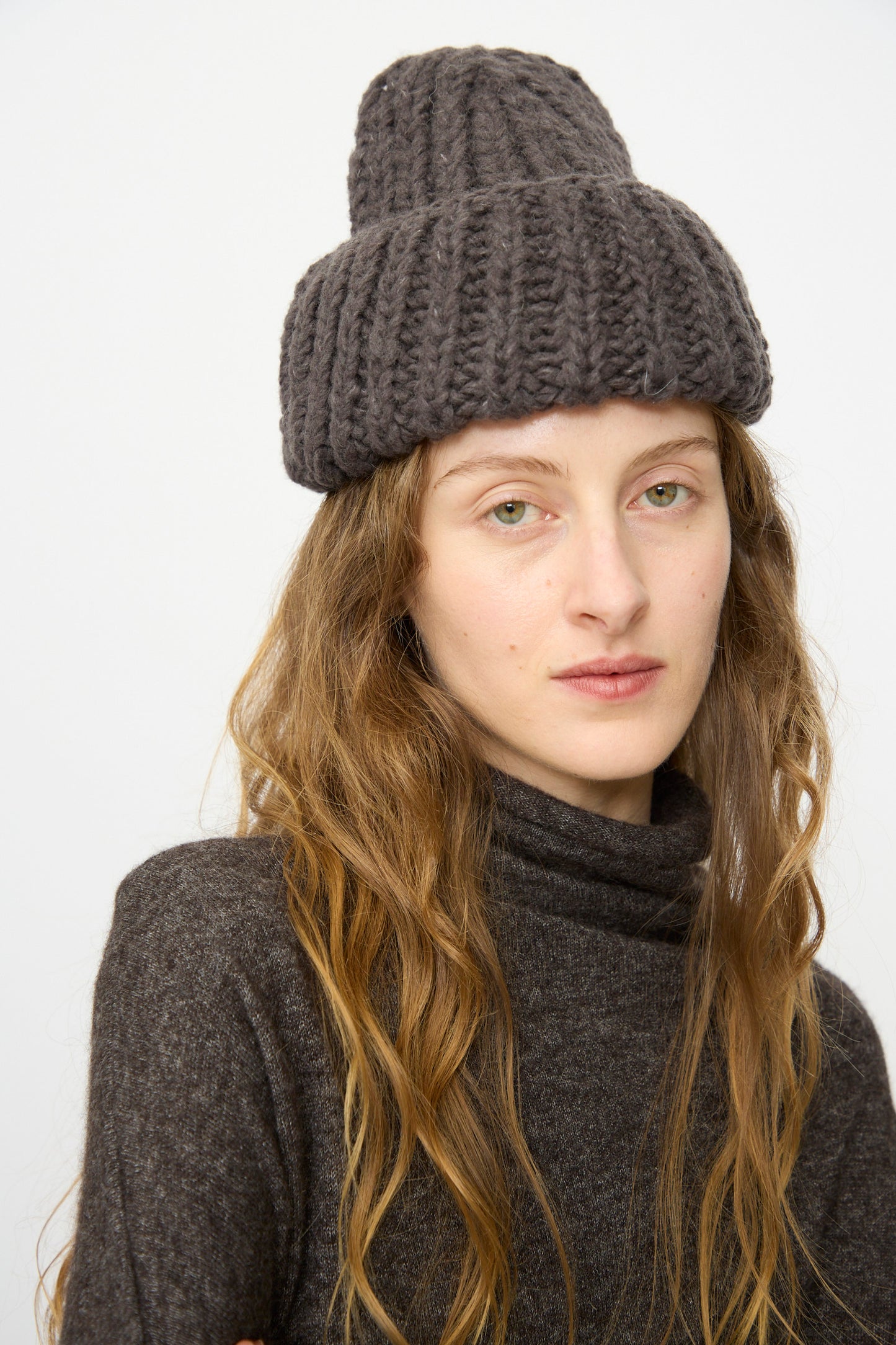 A person with long wavy hair wears the Lauren Manoogian Handknit Rib Hat in Coal—an oversized chunky gray merino wool hat—paired with a dark turtleneck sweater against a plain background.