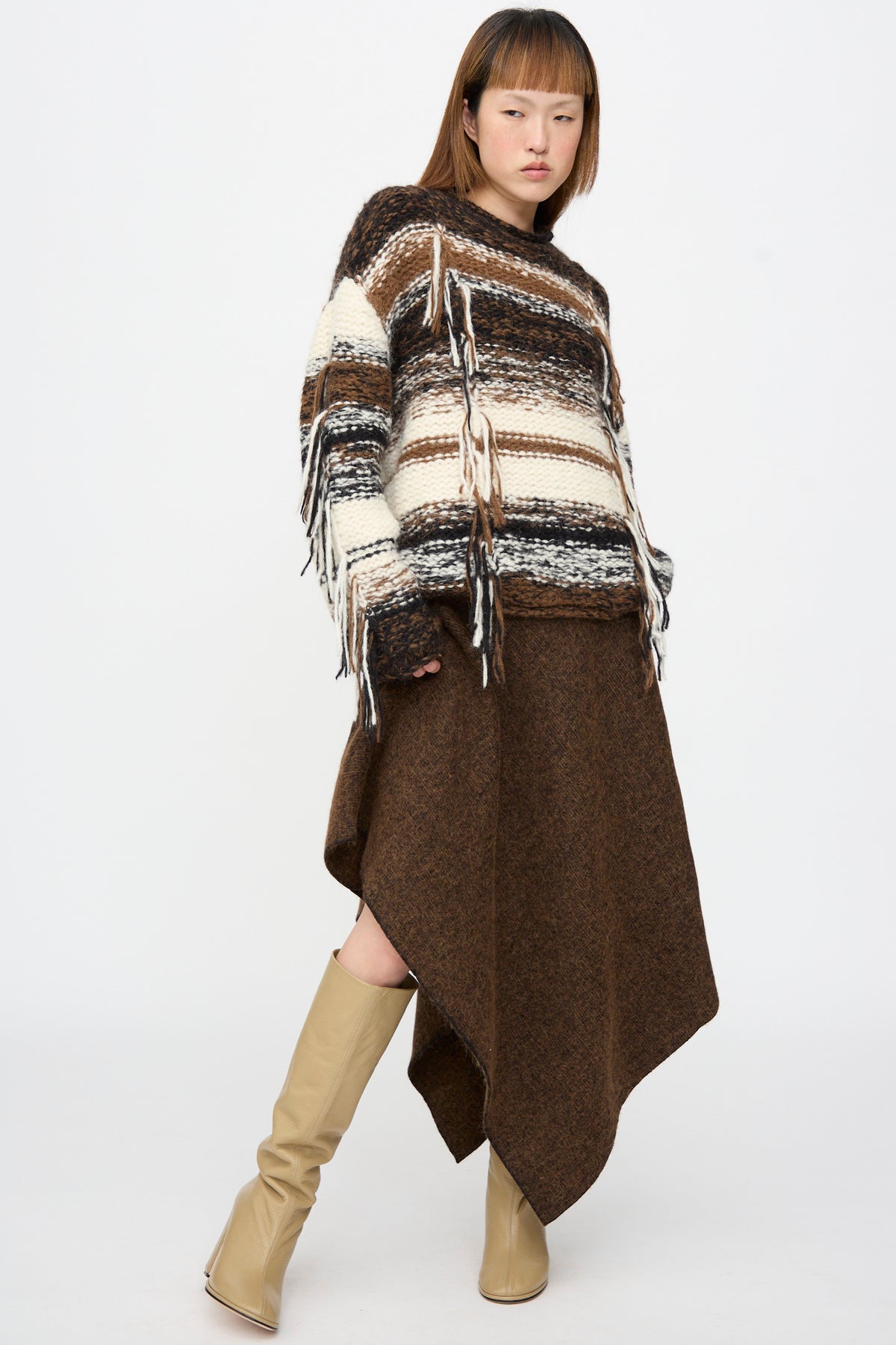 A person wearing a Lauren Manoogian Handknit Rio Rollneck in Marl Stripe with fringe, an asymmetrical brown skirt, and tan knee-high boots.