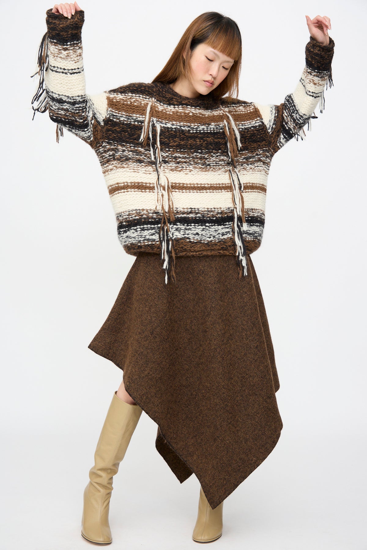 A person wearing a Lauren Manoogian Handknit Rio Rollneck in Marl Stripe with fringe details, an asymmetrical brown skirt, and beige knee-high boots.