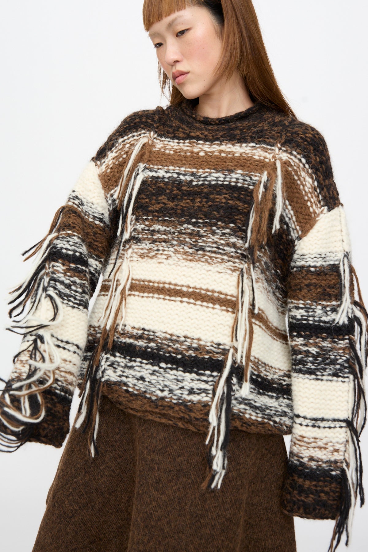 A person wears a Lauren Manoogian Handknit Rio Rollneck in Marl Stripe, an oversized, striped alpaca knit sweater featuring fringe details along the front and sleeves.