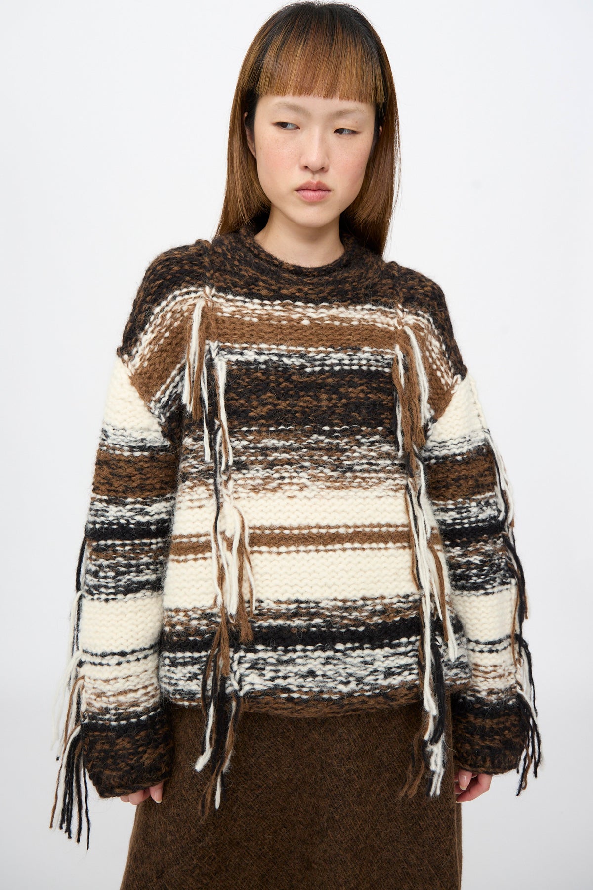 A person wears a Lauren Manoogian Handknit Rio Rollneck in Marl Stripe, an oversized alpaca knit sweater with brown, black, and cream stripes and vertical yarn fringe accents on the front and sleeves.