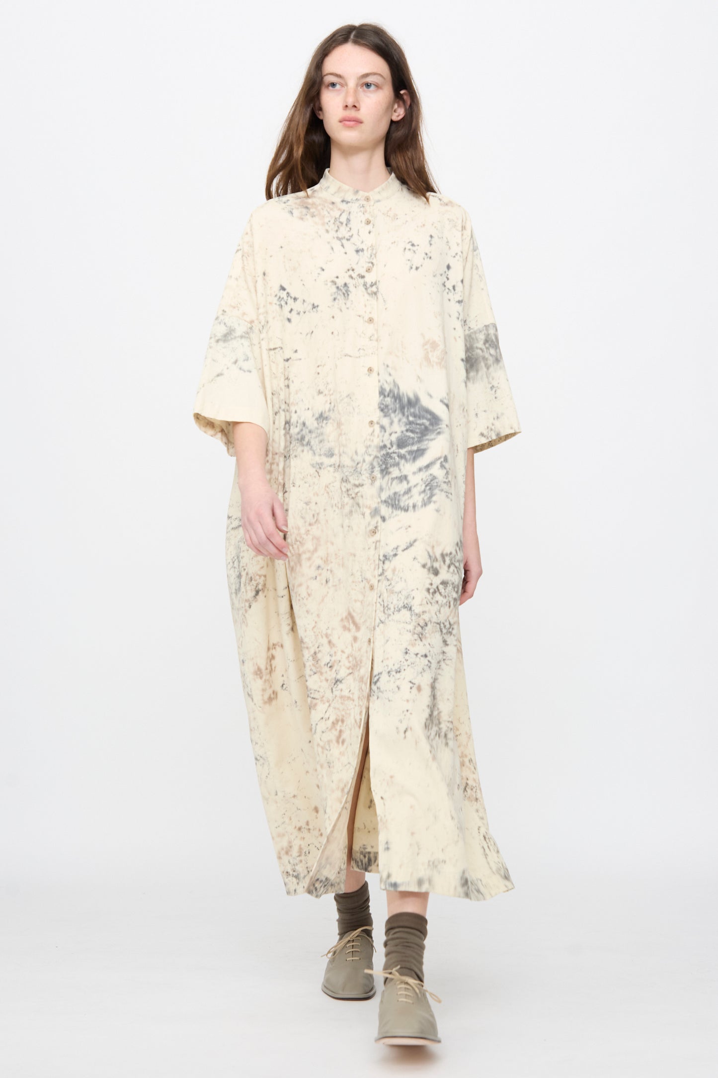 A person with mid-length brown hair stands against a light background, wearing the Lauren Manoogian Handpaint Shirt Dress in Natural with gray and brown tie-dye patterns and beige shoes.