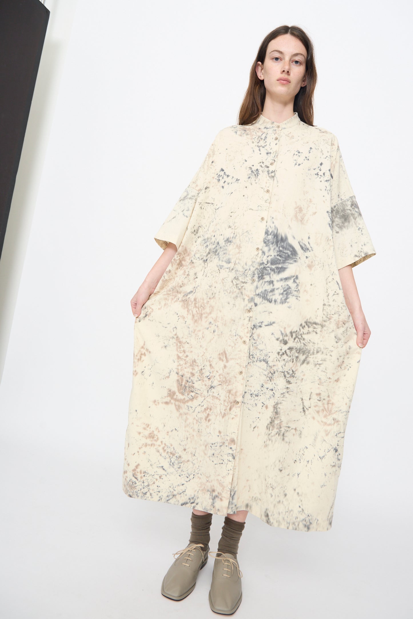 A woman stands against a white background wearing the Lauren Manoogian Handpaint Shirt Dress in Natural, a loose, ankle-length cream pima cotton dress with abstract grey and beige patterns, paired with brown socks and beige lace-up shoes. Made in Peru.
