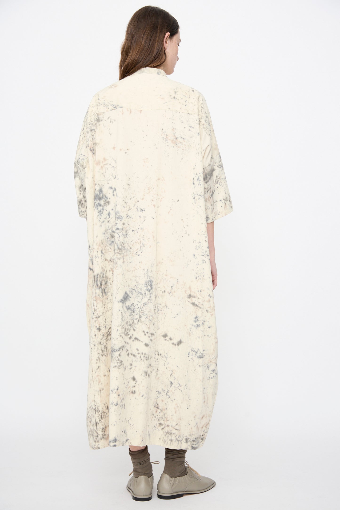 Person with their back to the camera wears the Lauren Manoogian Handpaint Shirt Dress in Natural, featuring beige and gray splatters, paired with gray socks and light gray shoes against a plain white background.
