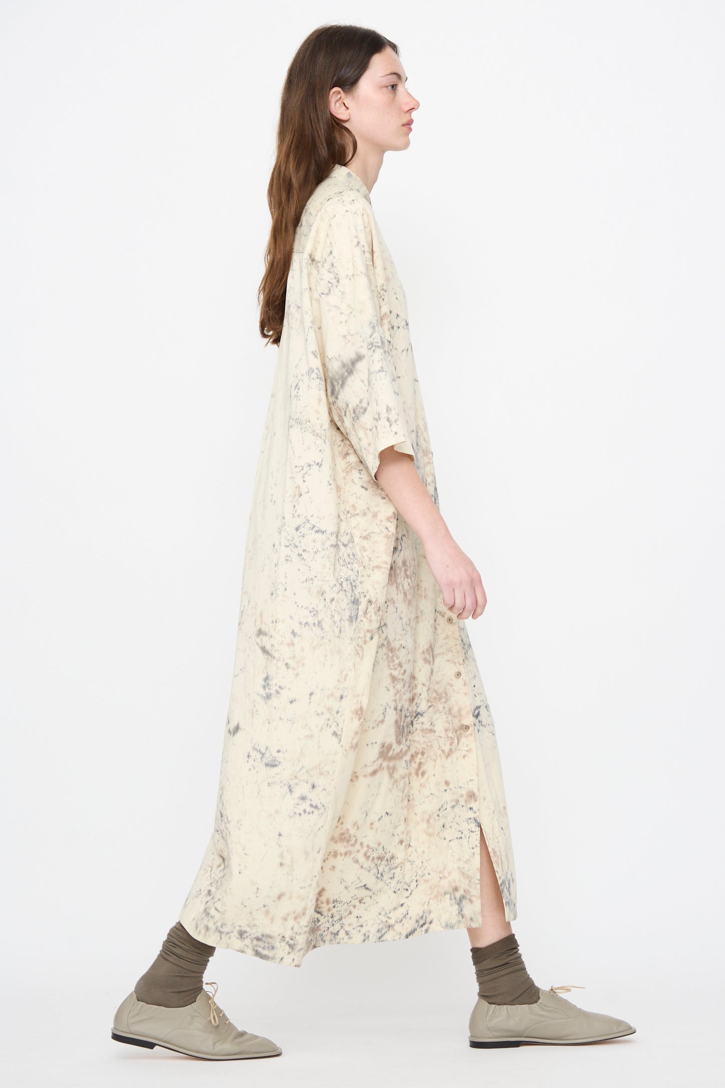 A woman in the Lauren Manoogian Handpaint Shirt Dress in Natural, a cream-colored long pima cotton piece with a marbled pattern, walks against a white background wearing taupe socks and shoes. 