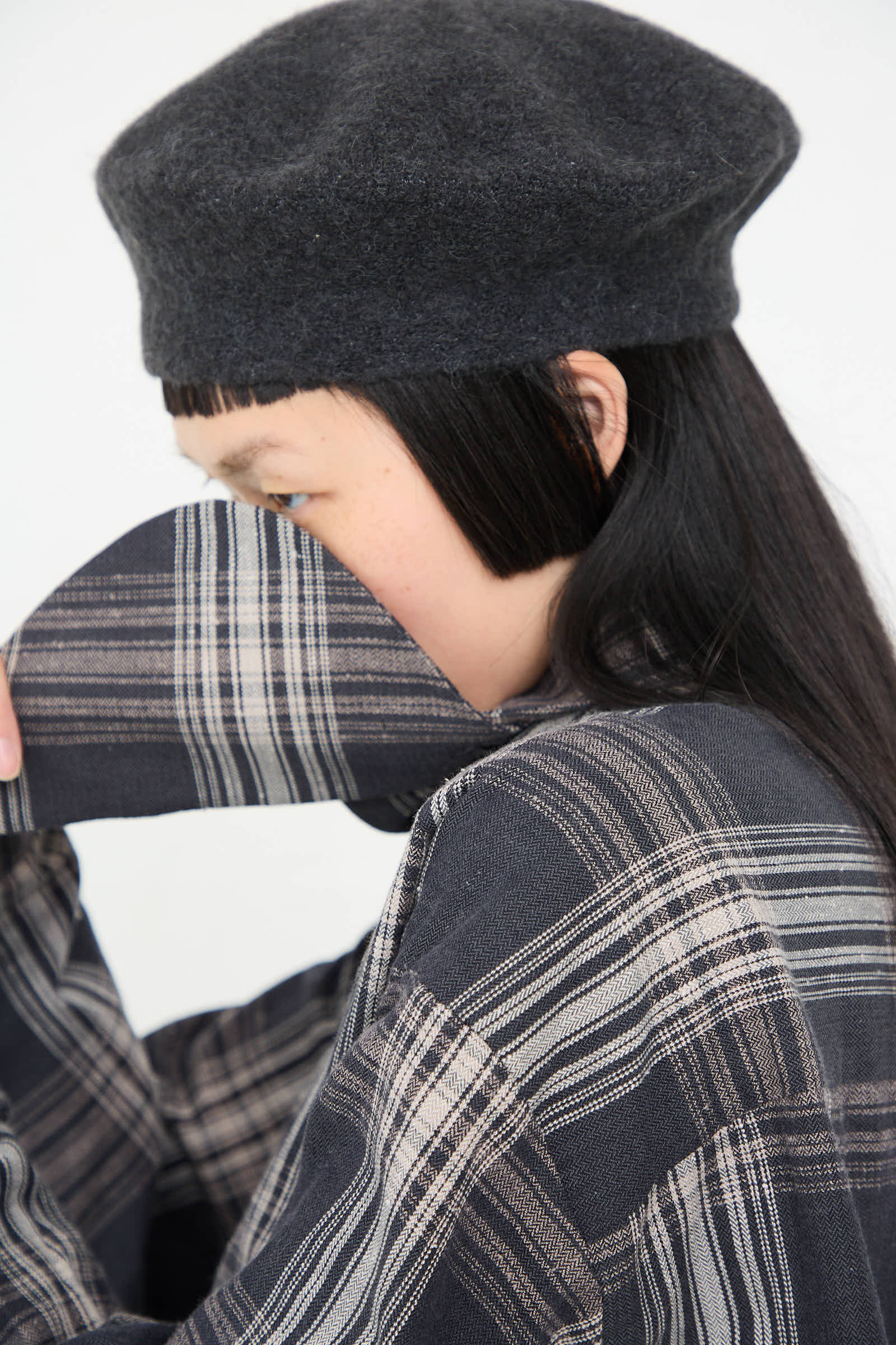 A person wearing a Horizontal Beret in Ink by Lauren Manoogian and plaid clothing, partially covering their face with their sleeve.