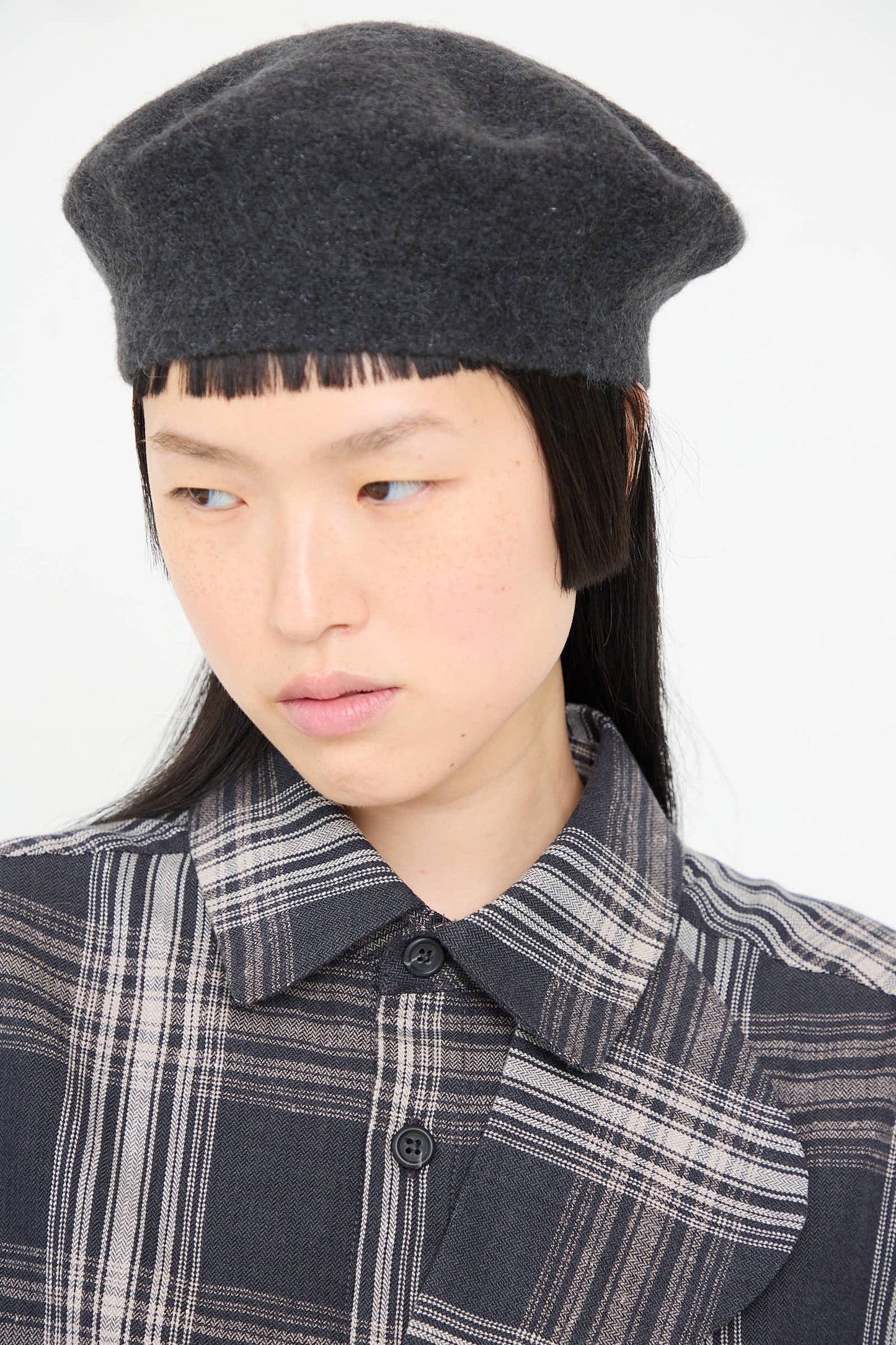 A person with straight dark hair wearing the Horizontal Beret in Ink by Lauren Manoogian, made in Peru from baby alpaca, and a plaid jacket, is looking down and to the side against a plain background.