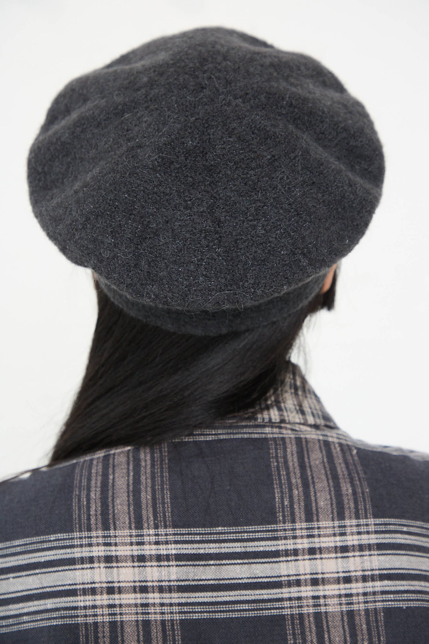 A person with long black hair is wearing the Lauren Manoogian Horizontal Beret in Ink and a plaid shirt, seen from the back.