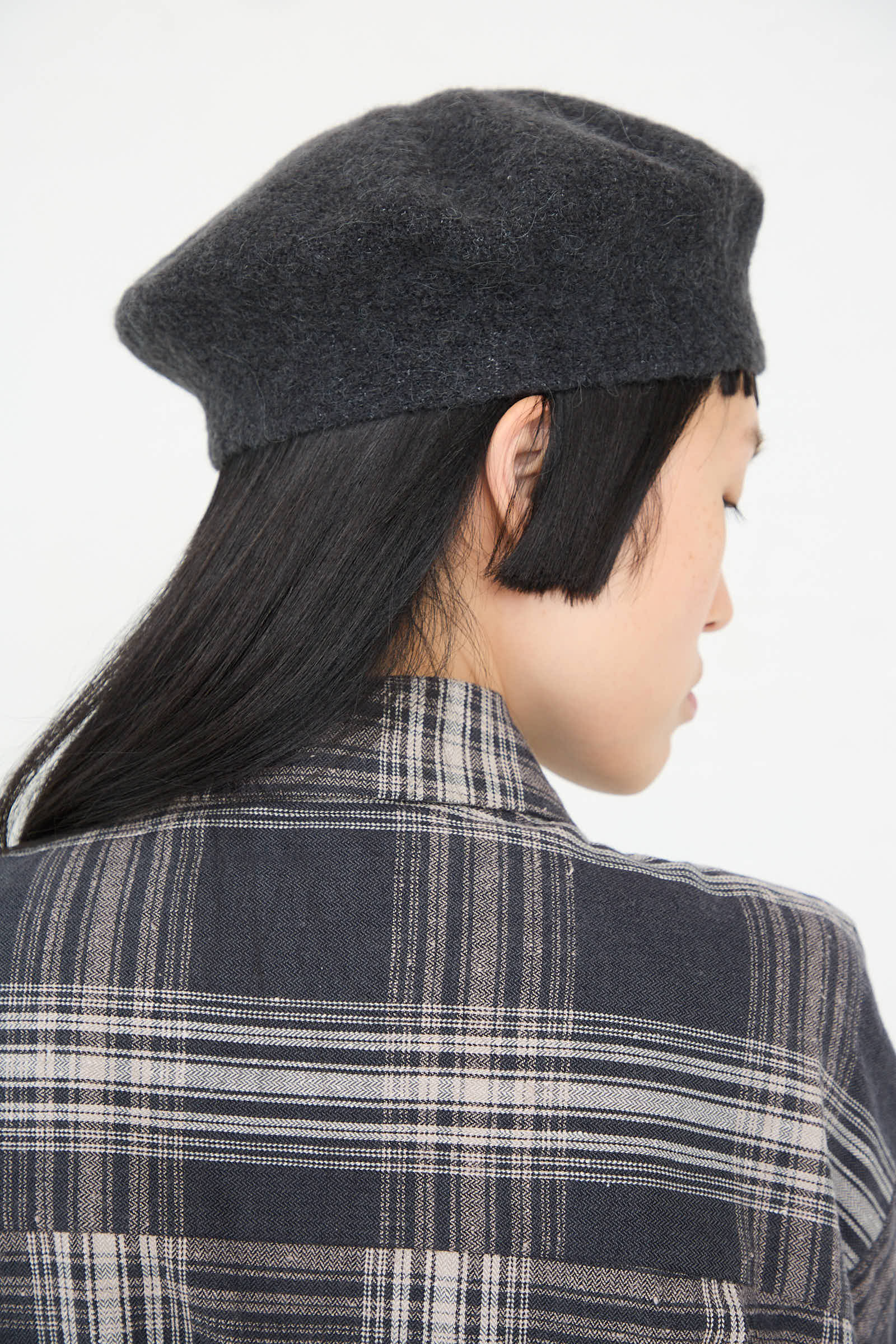 A person with long black hair and short bangs is shown from behind. They are wearing the Lauren Manoogian Horizontal Beret in Ink, made of baby alpaca wool, and a plaid shirt.