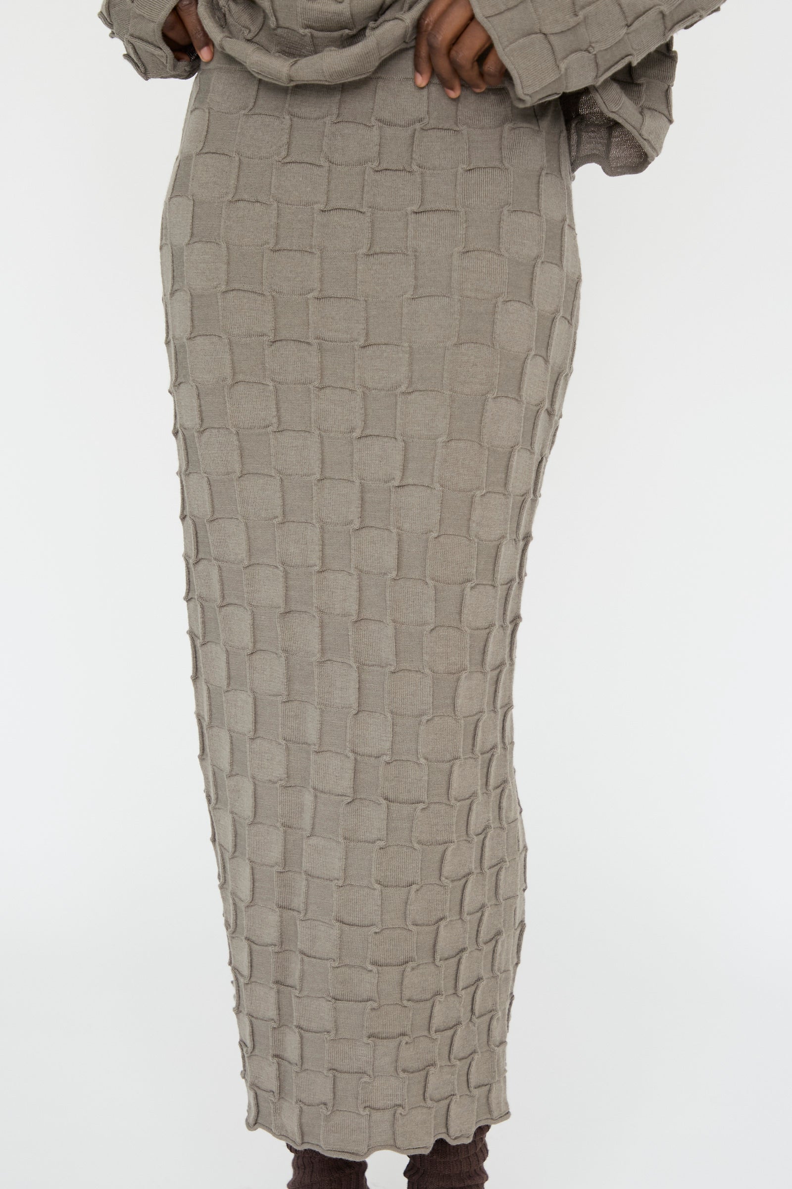 A person showcases the Lauren Manoogian Inlay Skirt in Concrete, a long textured beige skirt made from soft merino wool with an elegant woven pattern, against a plain white background.