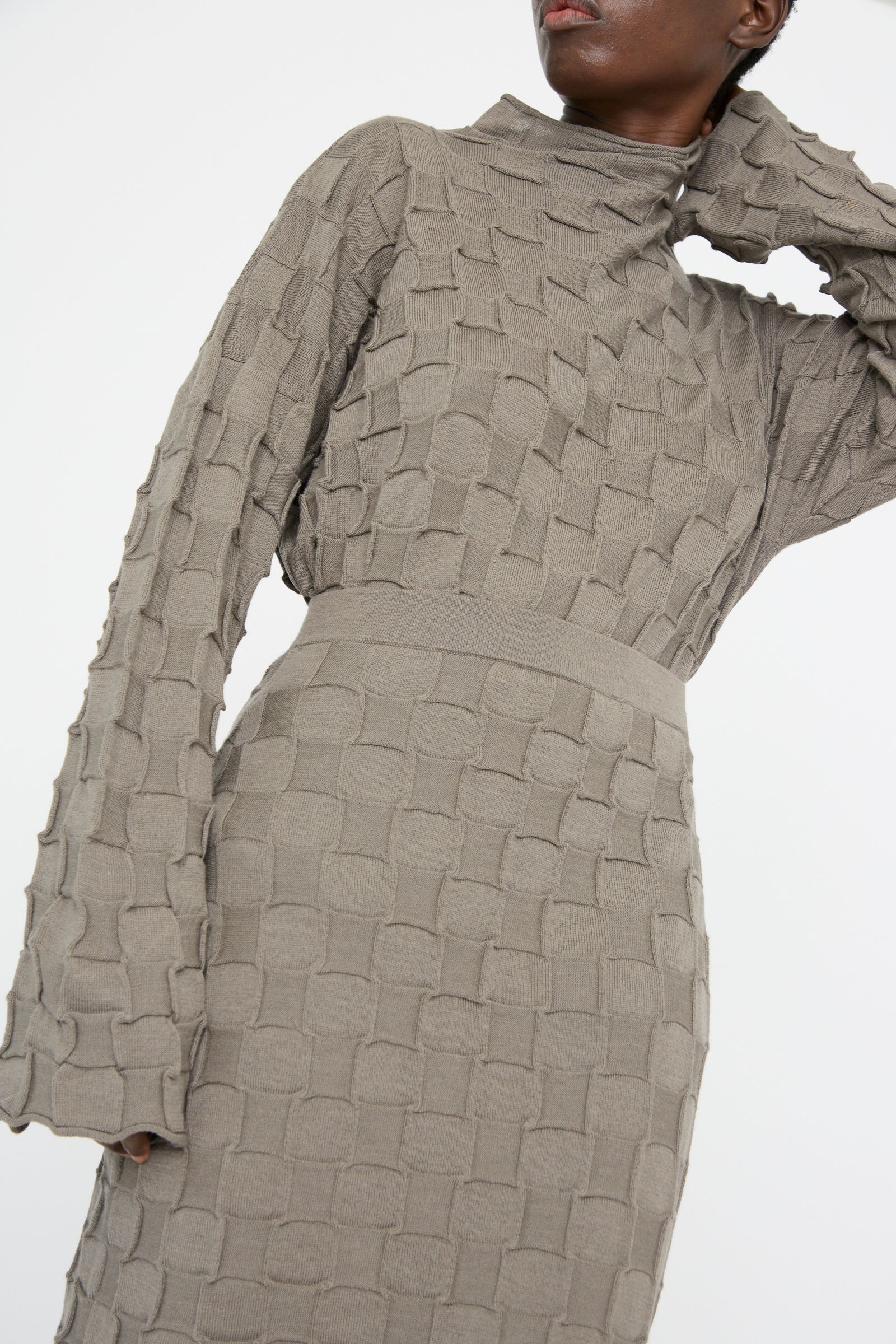 A person wearing a textured gray dress with long sleeves and a high neck, crafted from soft merino wool by Lauren Manoogian, similar in style to the Inlay Skirt in Concrete.