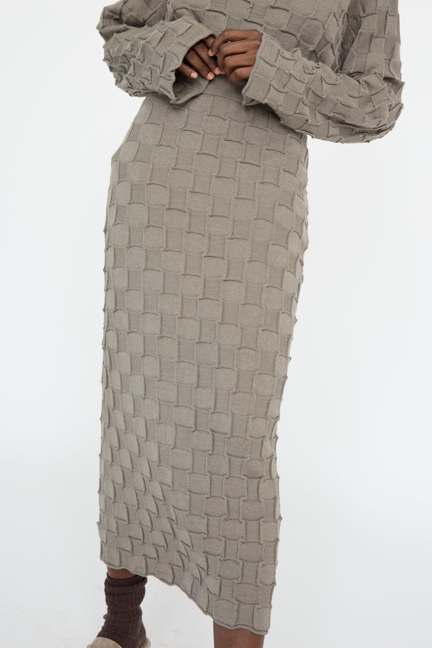 A person wears the Inlay Skirt in Concrete by Lauren Manoogian, a long, textured skirt made from merino wool with a woven pattern, against a plain white background.