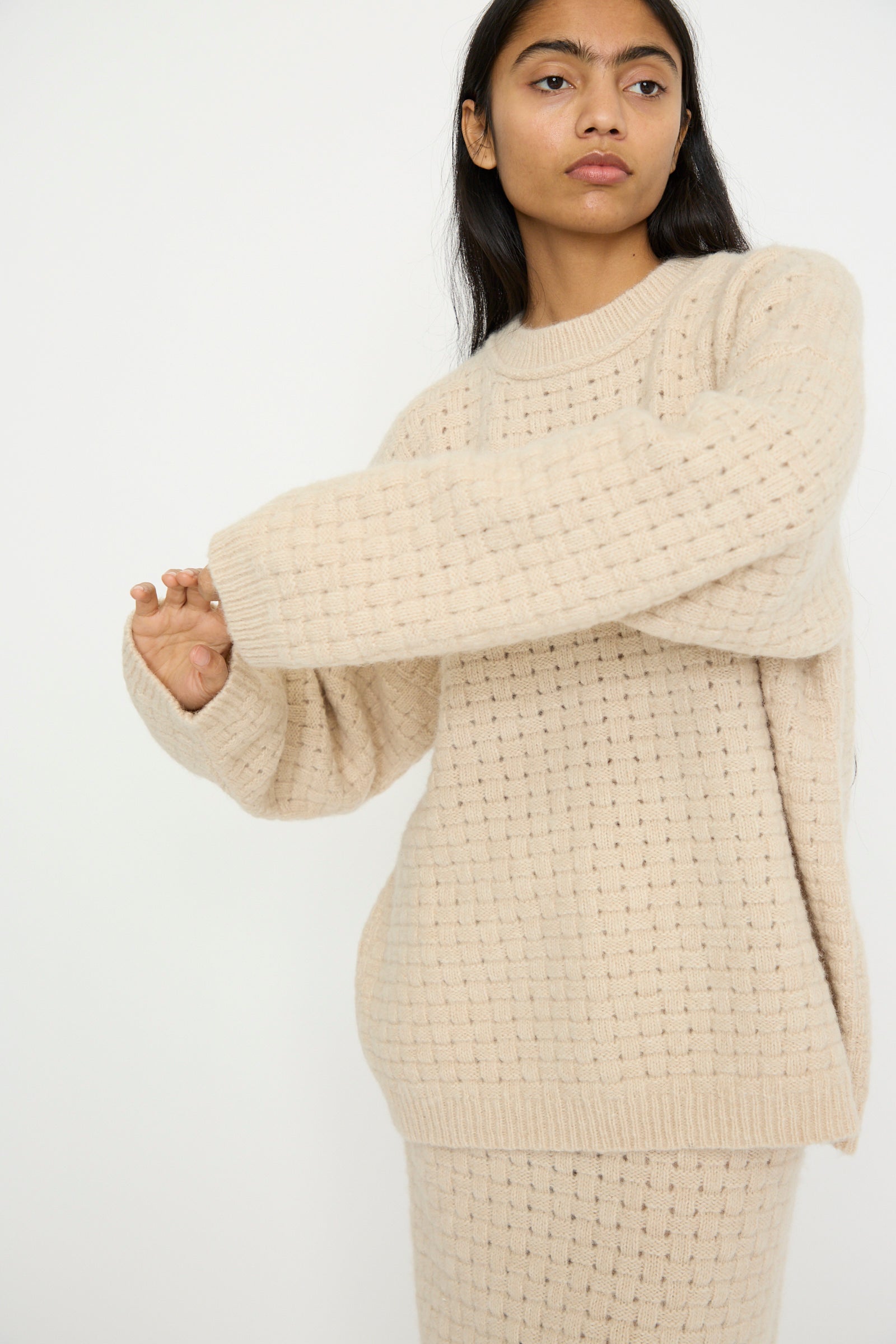 A person wearing the Lauren Manoogian Interlace Crewneck in Alabaster, crafted from an alpaca wool blend with a textured knit design, stands against a plain white background.