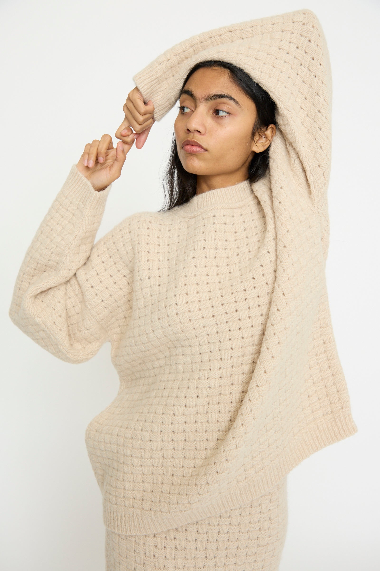 A person wearing the Lauren Manoogian Interlace Crewneck in Alabaster crafted from a cozy alpaca wool blend pulls one sleeve over their head while looking to the side.