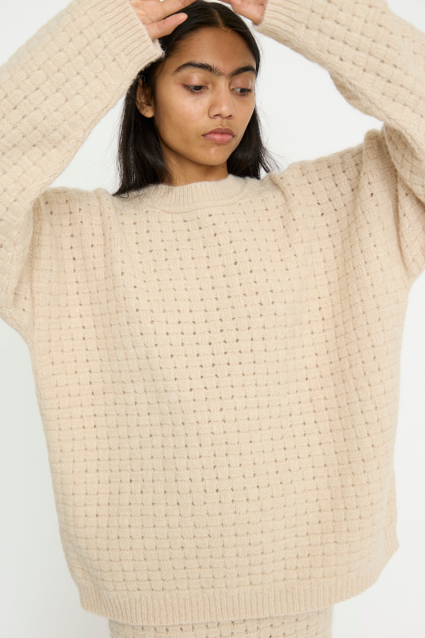 A person wearing the Lauren Manoogian Interlace Crewneck in Alabaster, an oversized knit sweater made from an alpaca wool blend, with arms raised against a plain background.