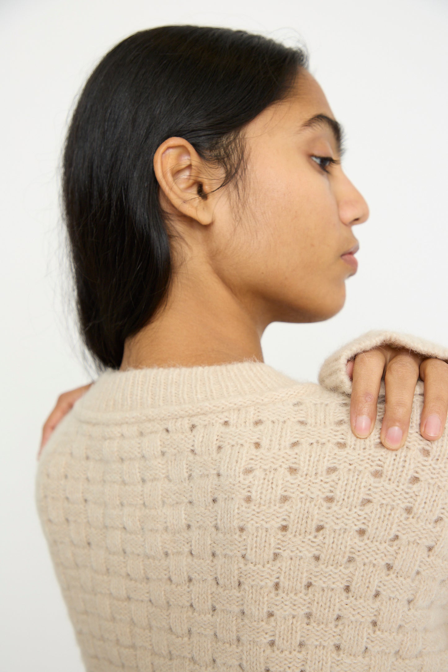 A person with long dark hair is seen from the side wearing the Lauren Manoogian Interlace Crewneck in Alabaster, an oversized textured beige sweater made from an alpaca wool blend.