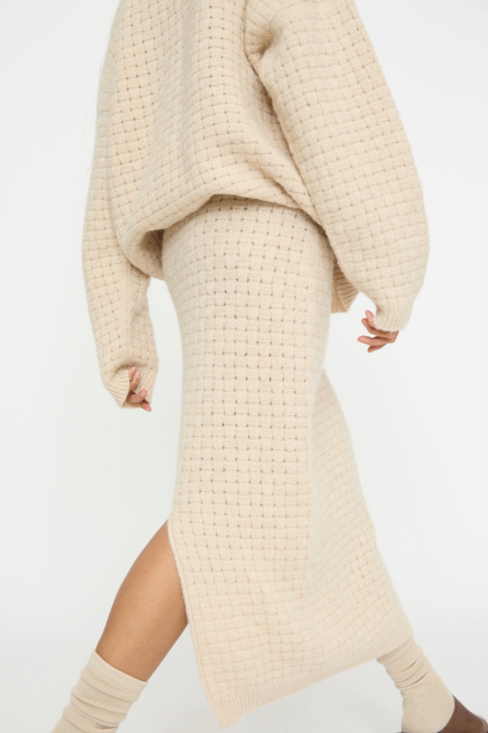 Dressed in a beige knit sweater and the Lauren Manoogian Interlace Skirt in Alabaster, handmade in Peru, a person walks against a white background and is seen from below the shoulders. 