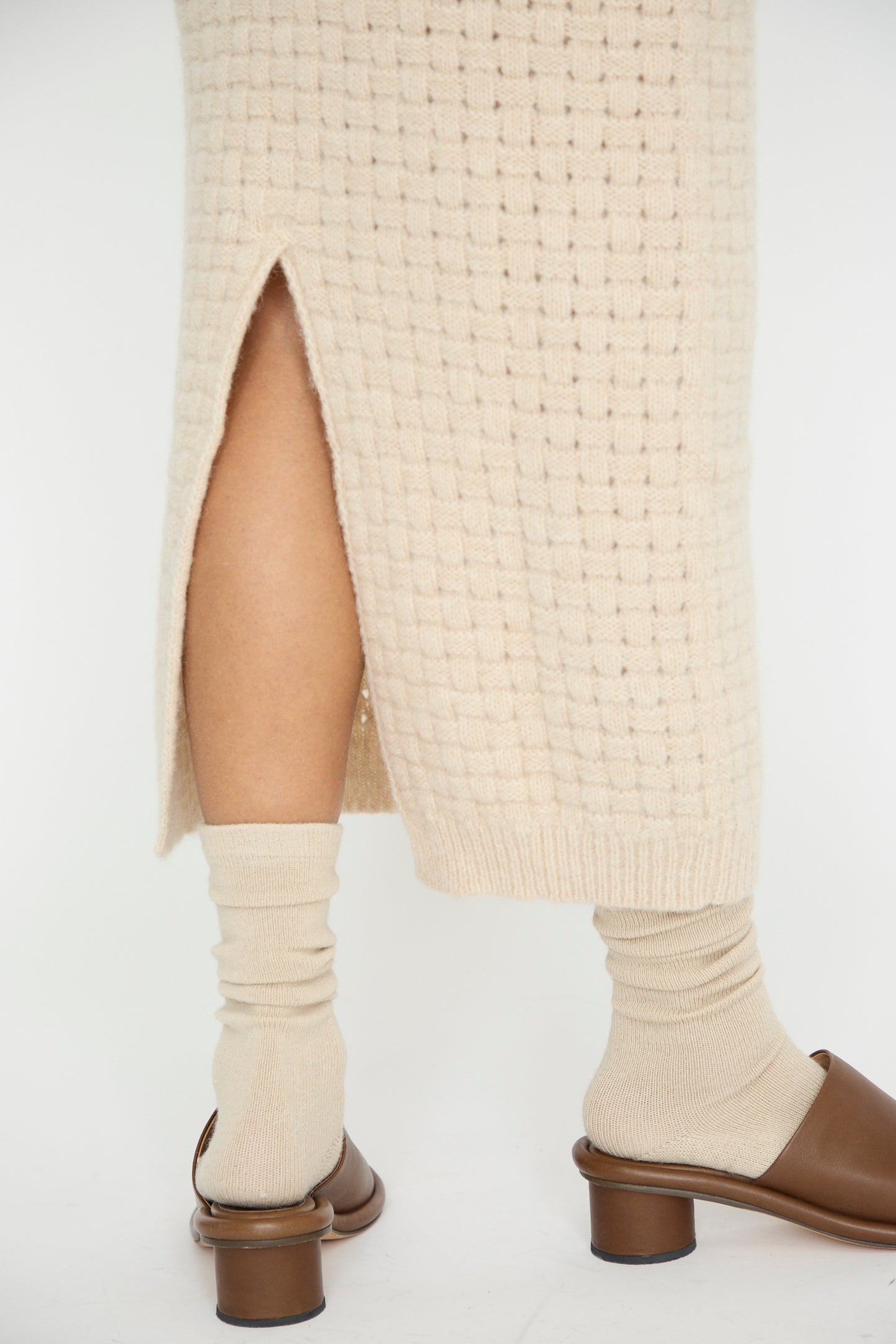 A person is facing away in Lauren Manoogian's Interlace Skirt in Alabaster, paired with beige socks and brown slide sandals. The image is cropped below the knees. 